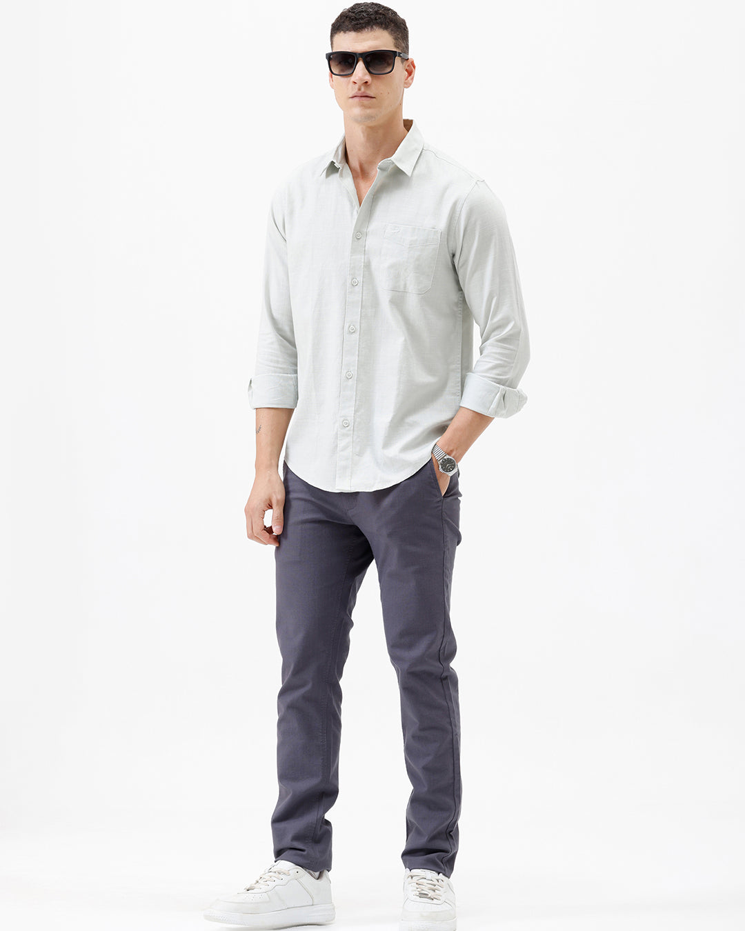 A man wearing a light sage green long-sleeve button-down shirt with rolled-up sleeves and dark gray trousers, standing against a plain white studio background.
