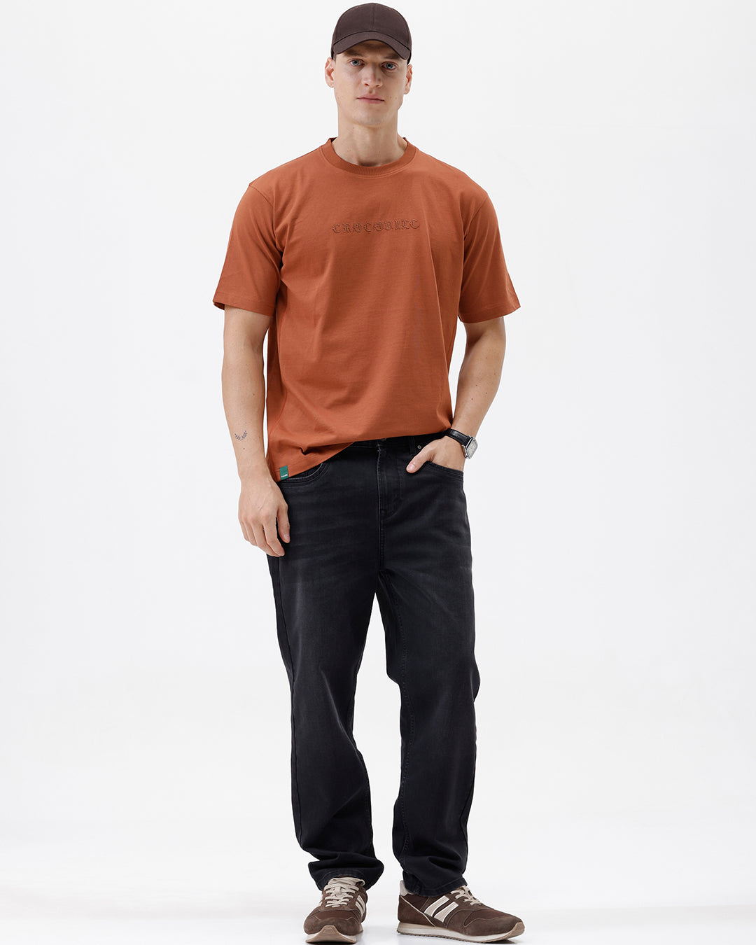 A man wearing a rust-orange crewneck t-shirt with a subtle tonal 'ELECTRIC' logo across the chest, paired with black pants, photographed against a clean white studio background.