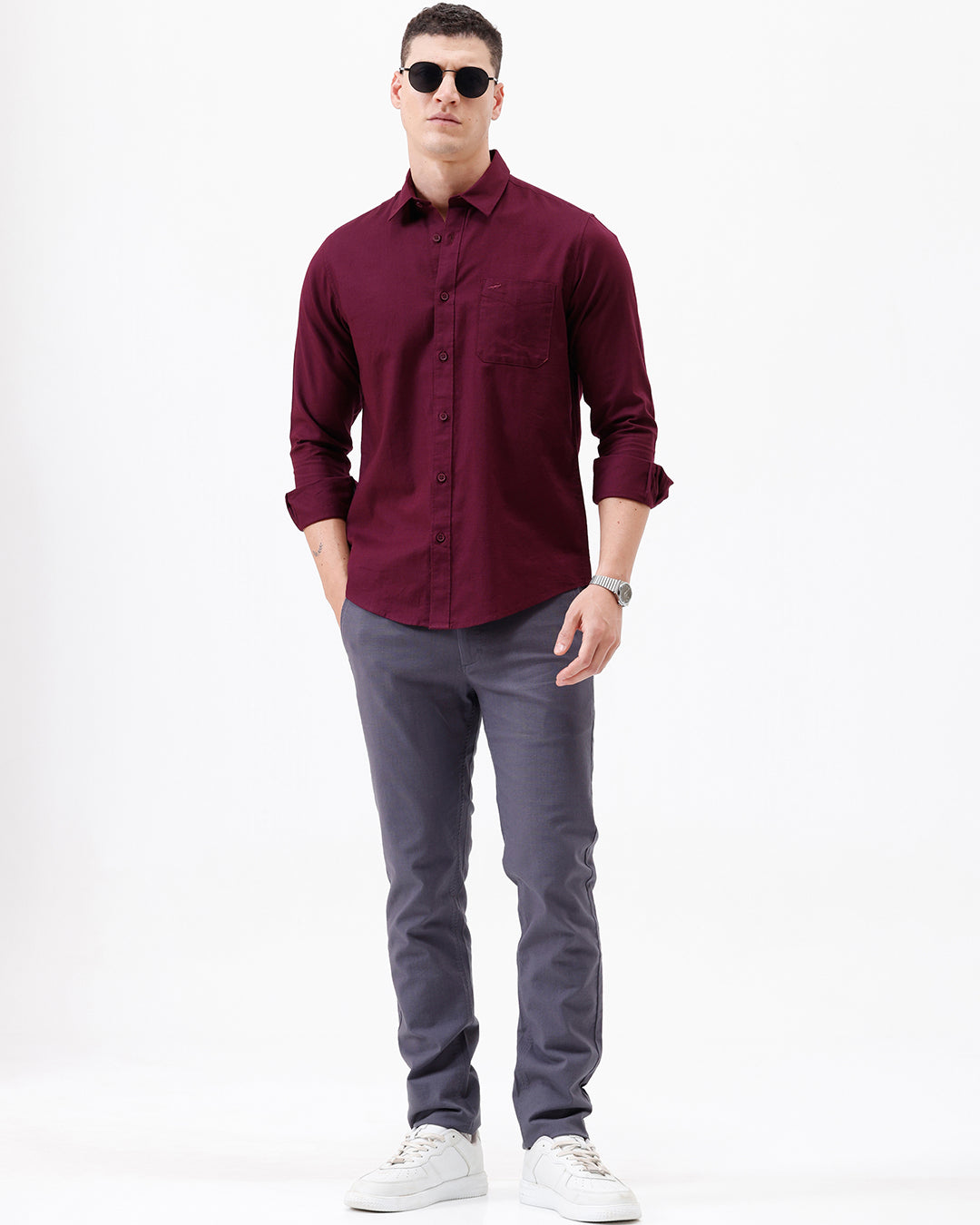 A man wearing a long-sleeve button-down shirt in deep burgundy with rolled-up sleeves and gray trousers, standing against a plain white studio background.