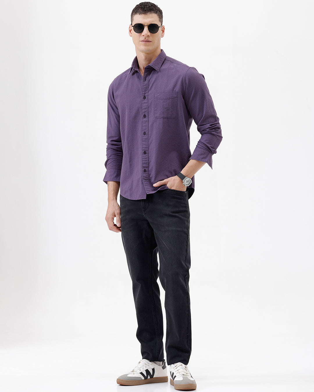 A man wearing a long-sleeve button-down shirt in a muted purple color, paired with black pants, standing against a plain white studio background.
