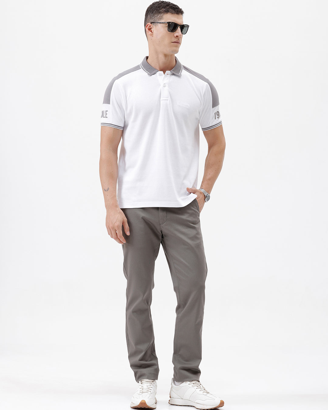 A man modeling a white short-sleeve polo shirt with gray shoulder panels and gray striped trim on the sleeves, paired with gray trousers and black sunglasses, against a plain white background.