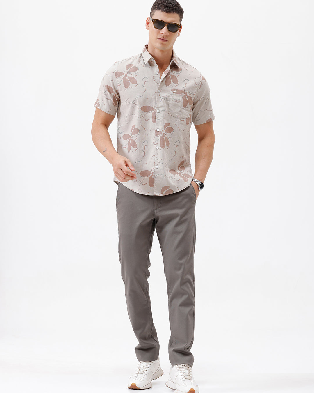 A man wearing a short-sleeve button-up shirt with a beige and brown floral pattern, paired with gray trousers, standing against a plain white background.