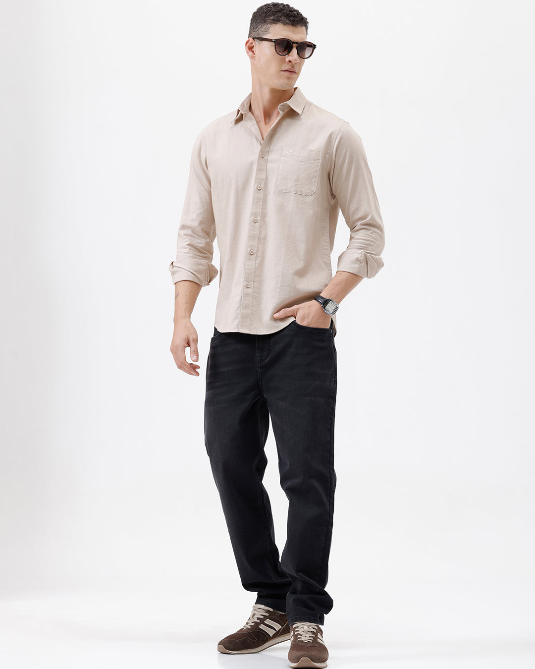 A man wearing a light beige long-sleeve button-up shirt with a chest pocket, paired with dark jeans, standing against a plain white studio background.