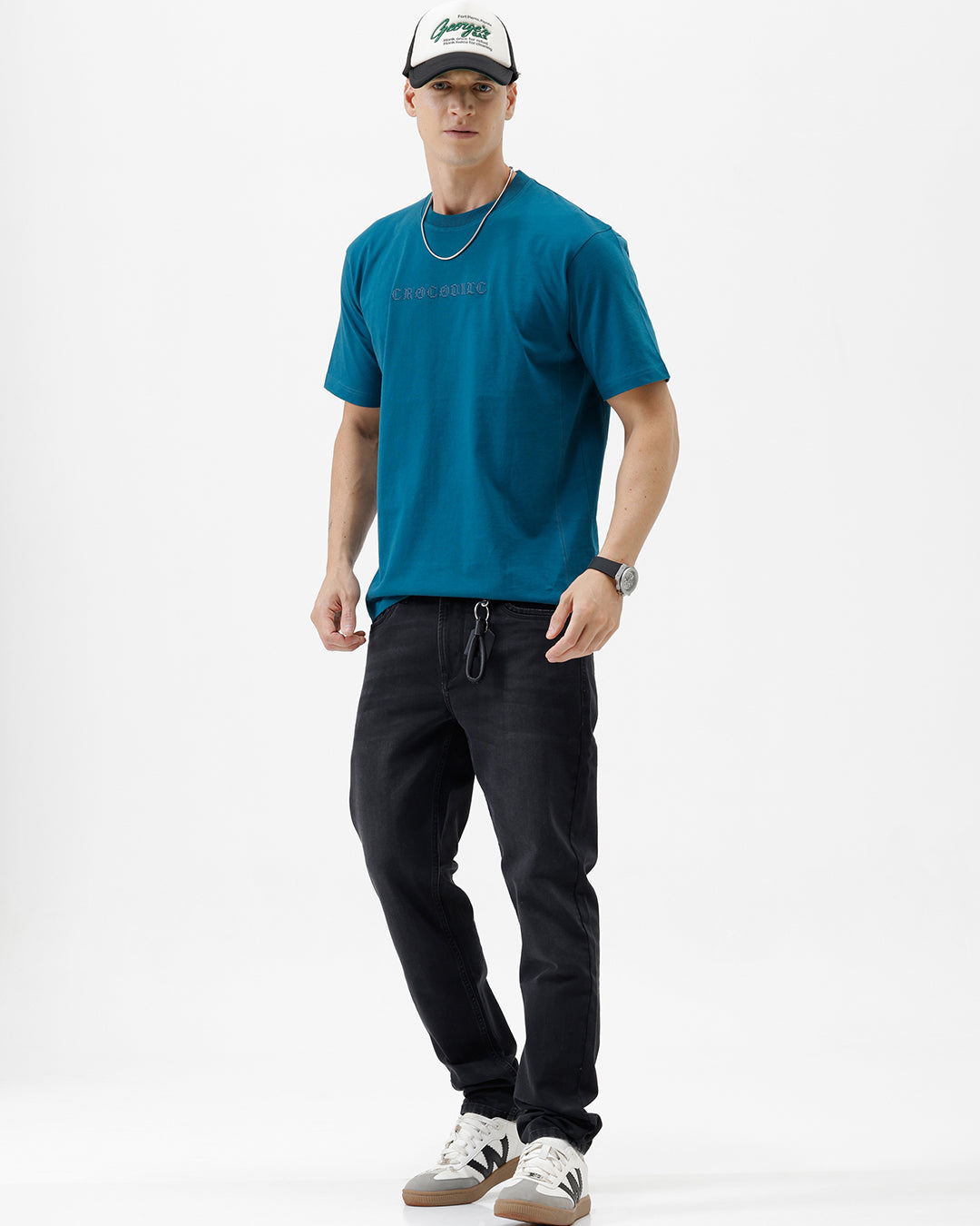 A man wearing a teal blue short-sleeve t-shirt with a subtle tonal 'GREATNESS' text graphic across the chest, paired with black pants, photographed against a clean white studio background.