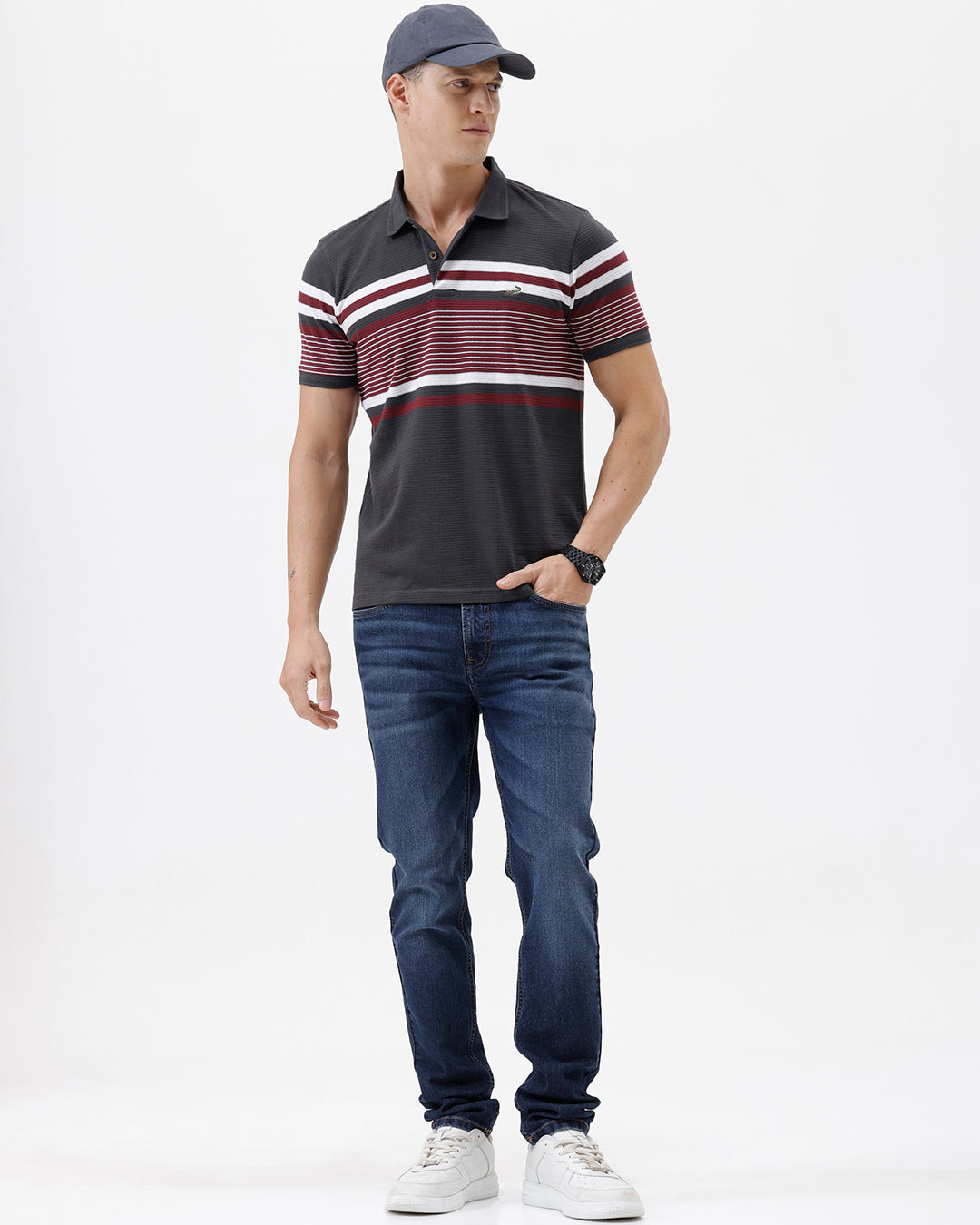 A man wearing a dark gray Lacoste polo shirt with horizontal red and white stripes across the chest and sleeves, paired with blue jeans, against a plain light gray background.