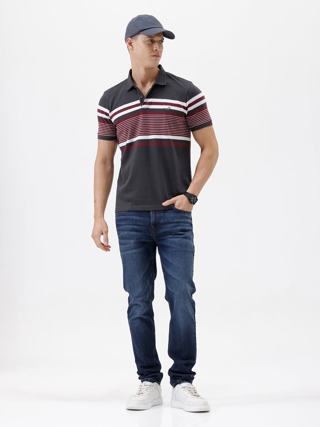 A man wearing a dark gray Lacoste polo shirt with horizontal red and white stripes across the chest and sleeves, paired with blue jeans, against a plain light gray background.