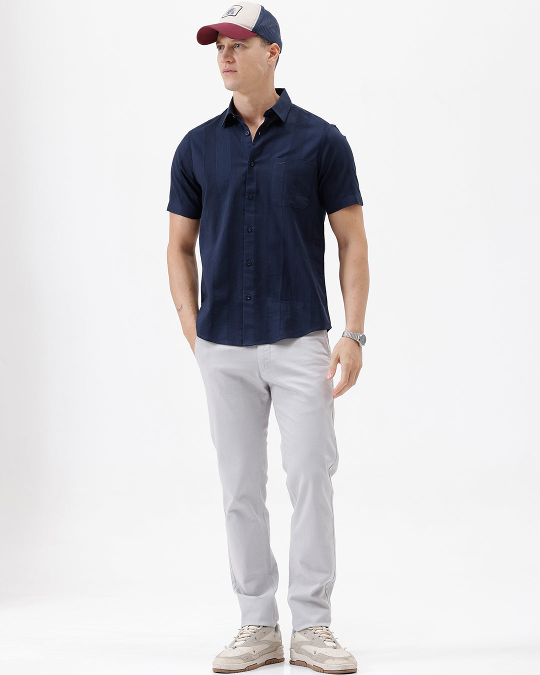 A man wearing a dark navy blue short-sleeve button-down shirt and light gray trousers, standing against a plain white studio background.