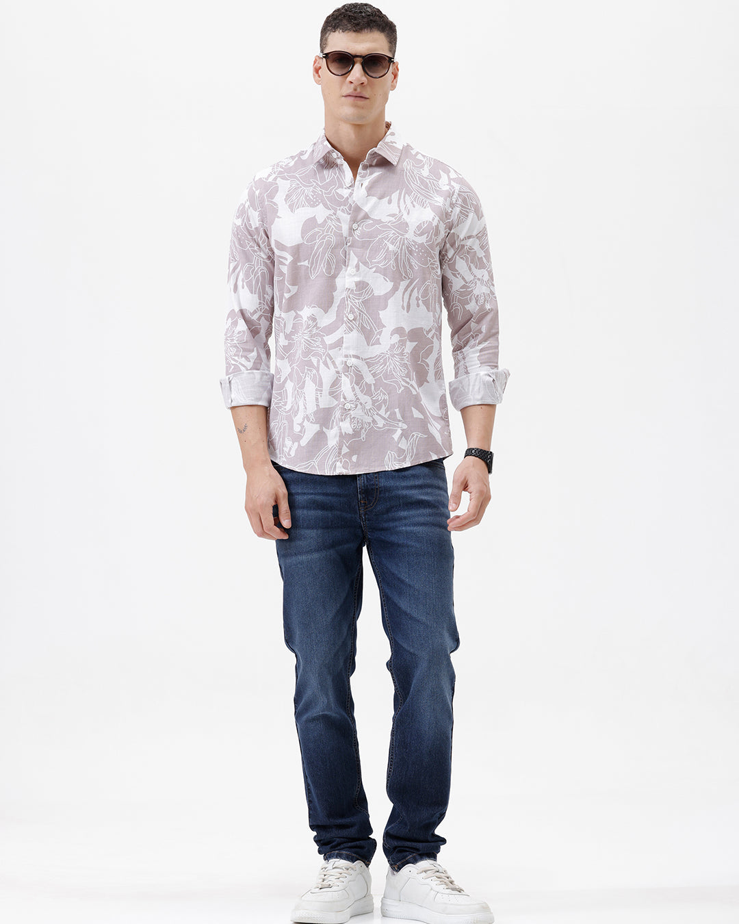 A man wearing a long-sleeve button-up shirt with a light gray and white abstract floral pattern, paired with dark blue jeans, standing against a plain white background.