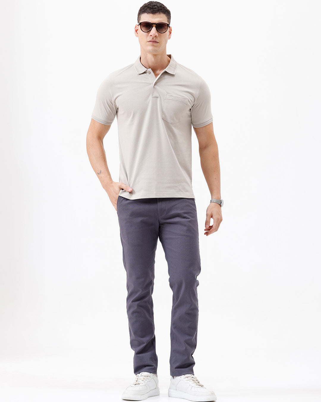 A man wearing a light beige short-sleeve polo shirt with a chest pocket, paired with dark gray trousers, standing against a plain white studio background.