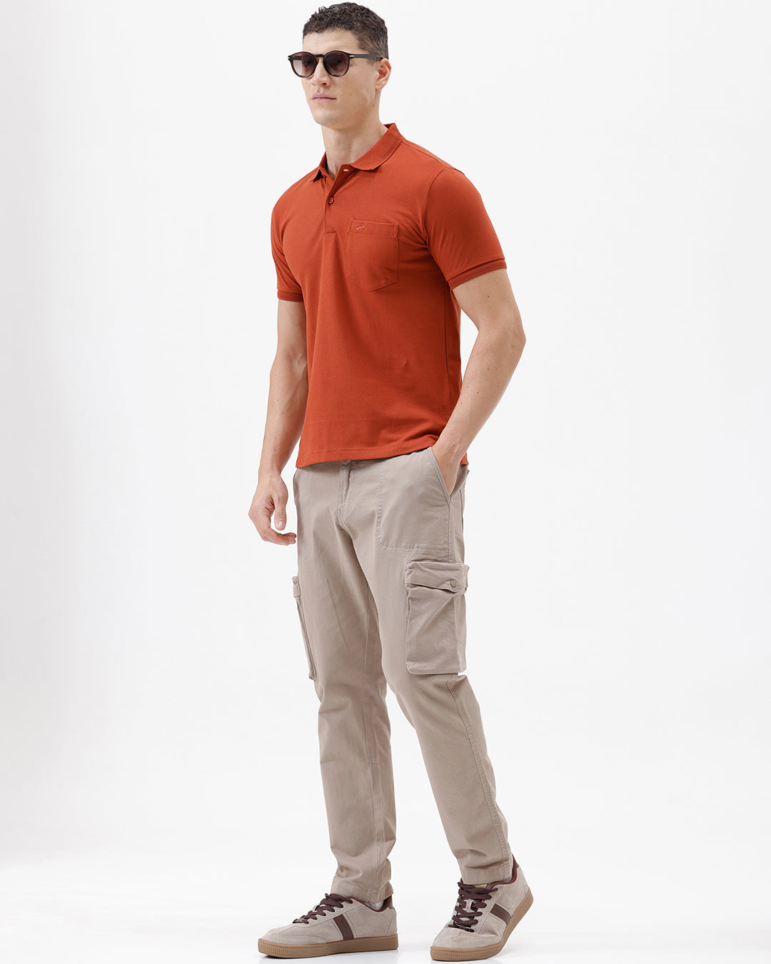A man wearing a rust-orange short-sleeve polo shirt with a chest pocket, paired with beige cargo pants, standing against a plain light gray studio background.