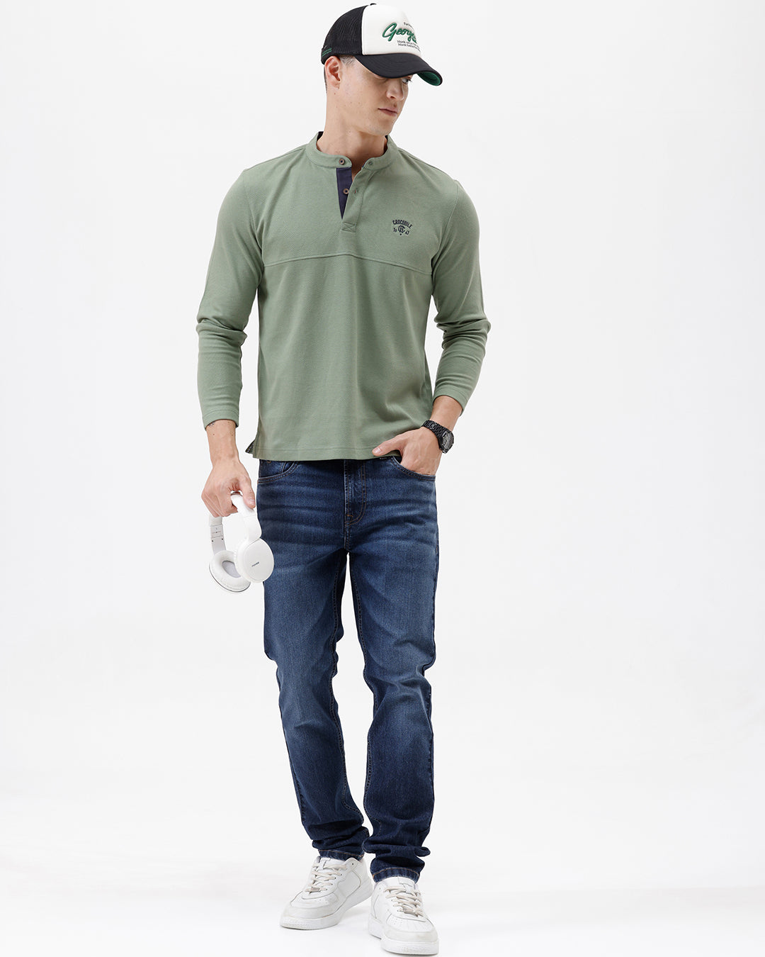 A man wearing a sage green long-sleeve henley shirt with a small embroidered logo on the left chest, paired with blue jeans, against a plain white studio background.