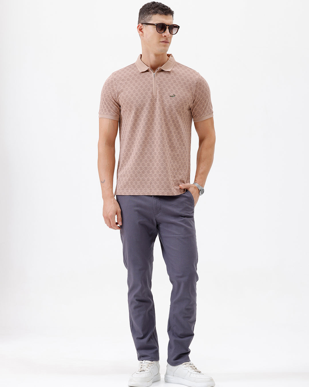 A man wearing a short-sleeve polo shirt with a muted brown and beige geometric pattern, paired with dark gray trousers, standing against a plain white studio background.