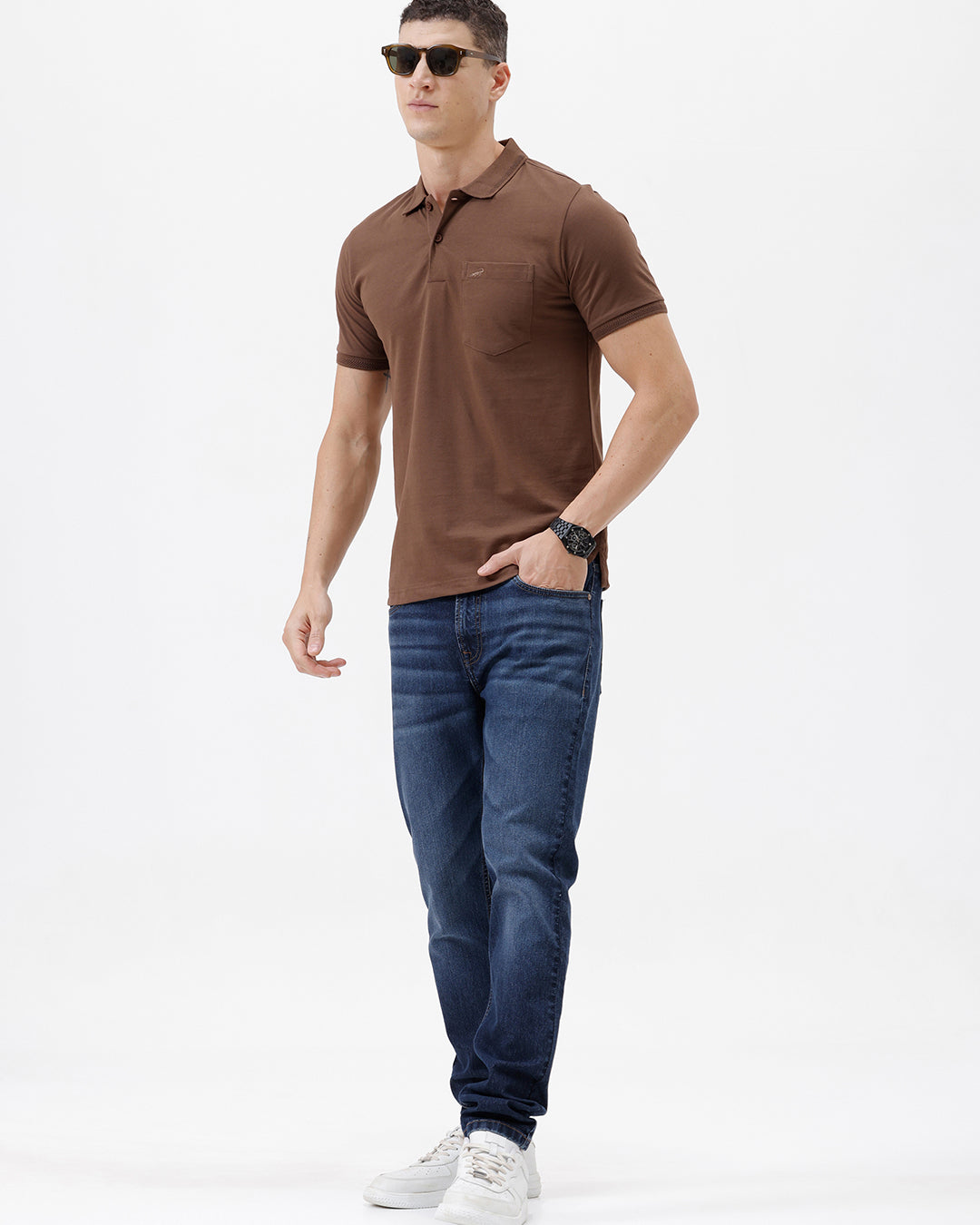 A man wearing a brown short-sleeve polo shirt with a chest pocket, paired with blue jeans, standing against a plain light gray background.