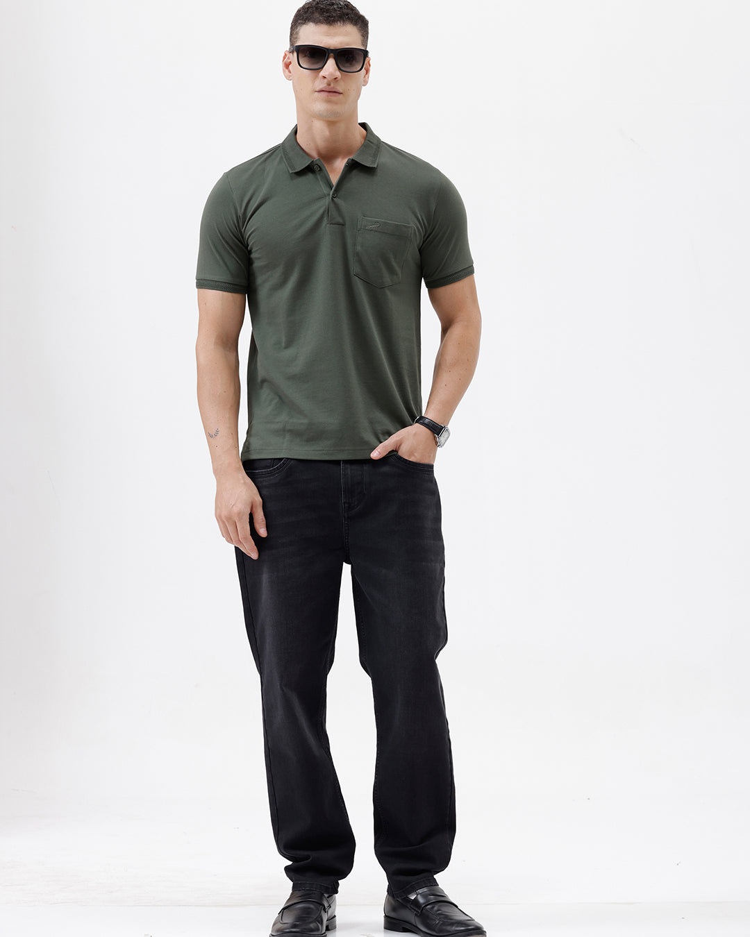 A man wearing a dark olive green short-sleeve polo shirt with a chest pocket, paired with dark jeans, standing against a plain light gray background.