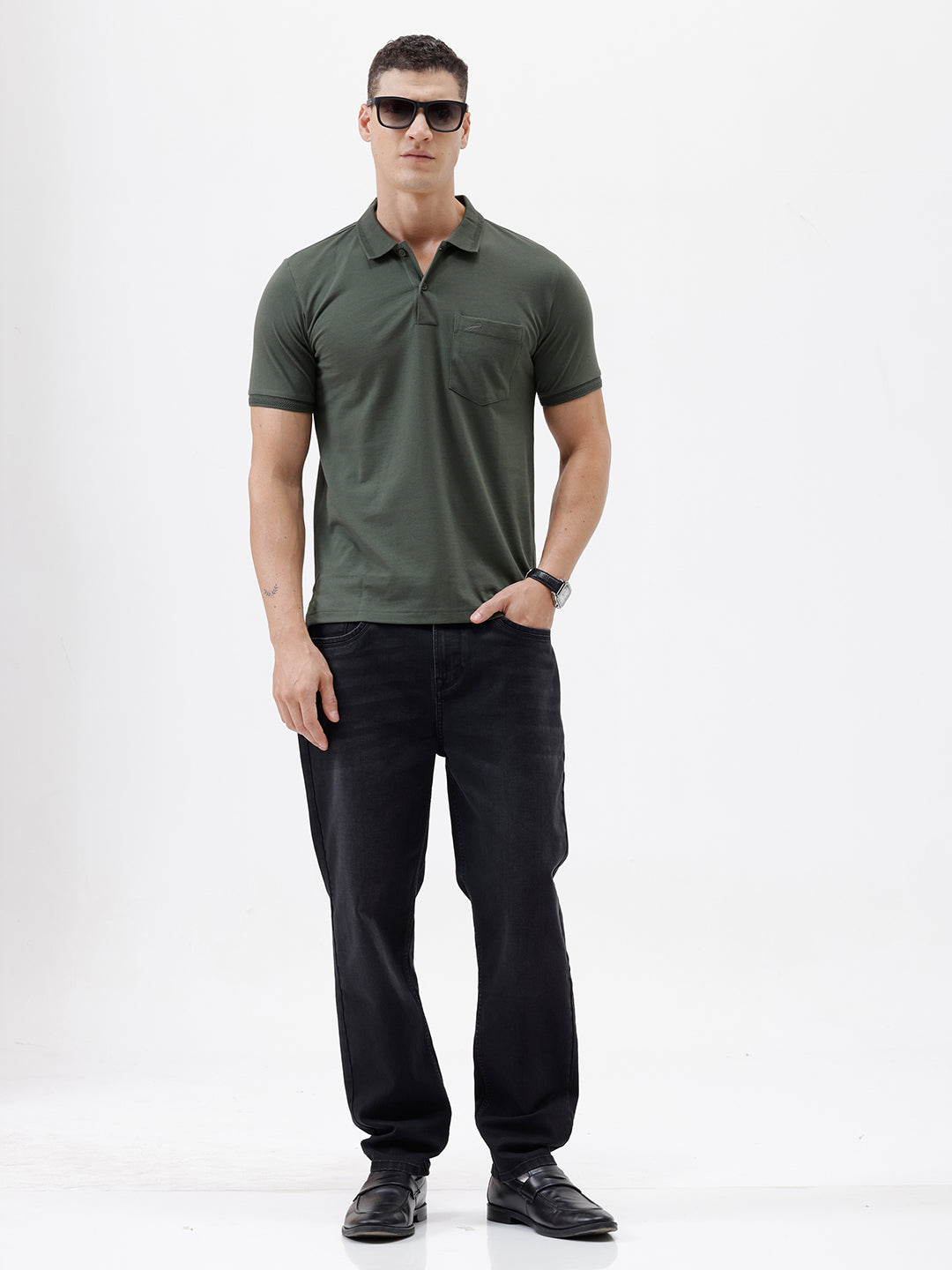 A man wearing a dark olive green short-sleeve polo shirt with a chest pocket, paired with dark jeans, standing against a plain light gray background.