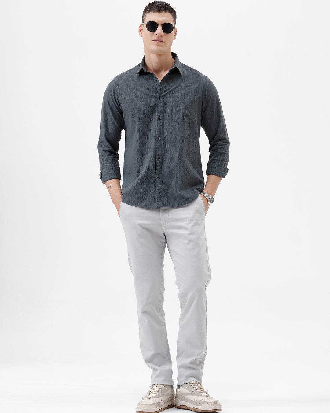 A man wearing a dark charcoal gray long-sleeve button-down shirt with a chest pocket, paired with light gray trousers, standing against a plain white studio background.