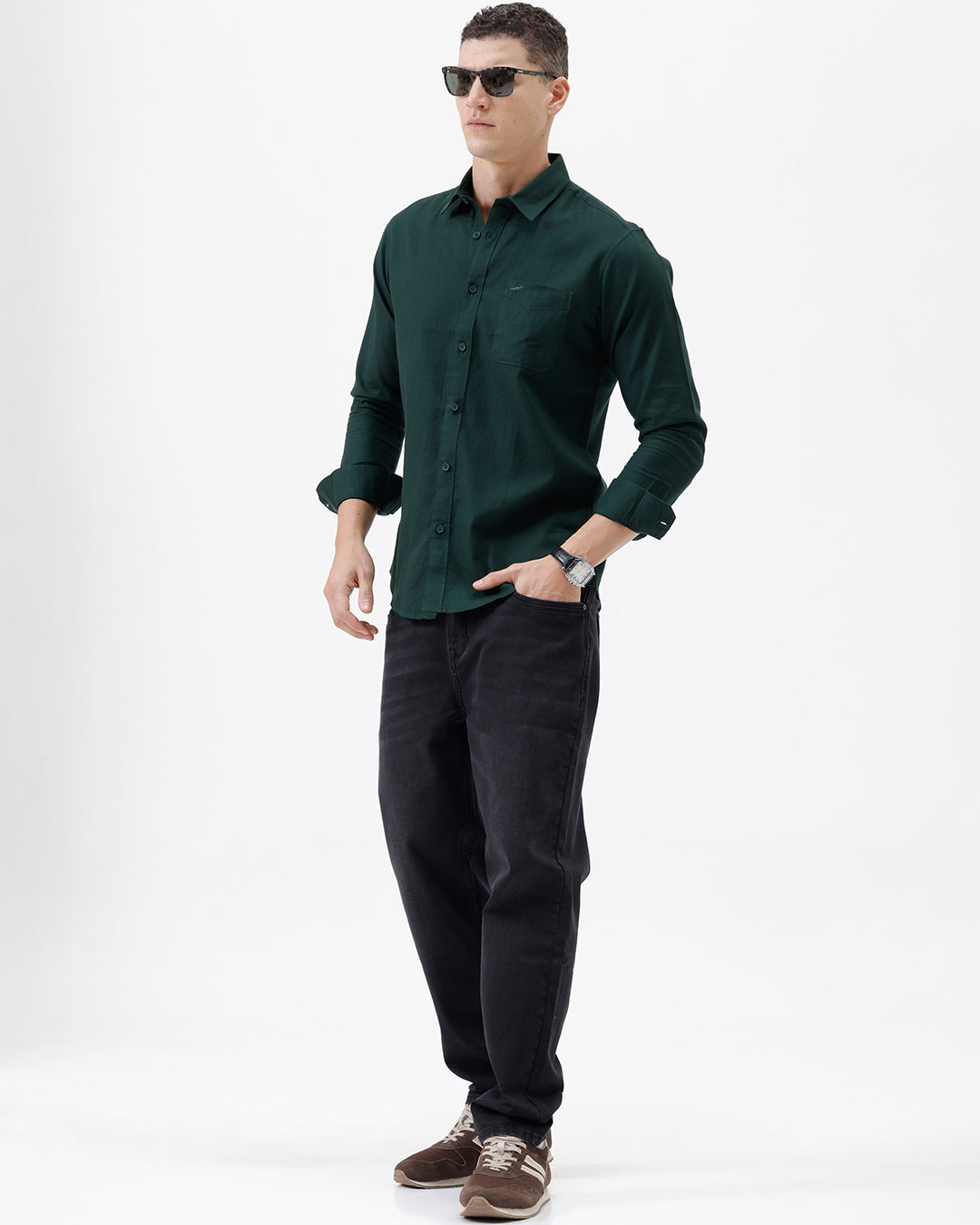 A man wearing a dark forest green long-sleeve button-down shirt with a chest pocket, paired with dark jeans, standing against a plain white studio background.