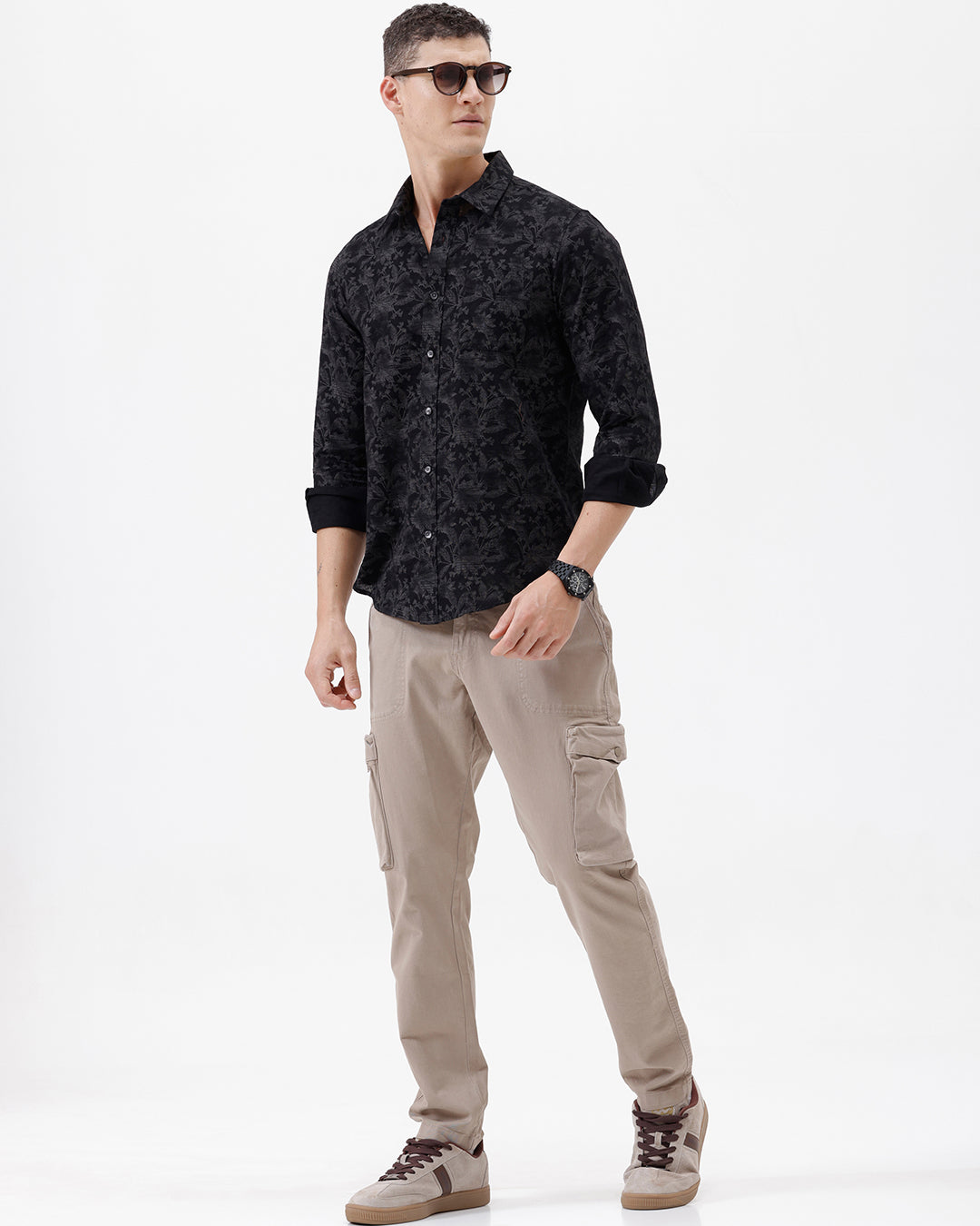 A man wearing a black long-sleeve button-up shirt with a subtle tonal floral pattern, paired with beige cargo pants and dark sunglasses, standing against a plain white background.