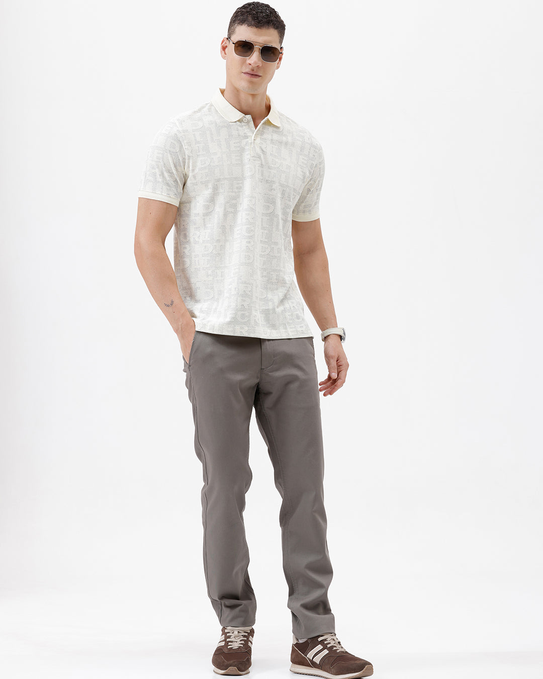 A man wearing a short-sleeve polo shirt with a subtle repeating monogram pattern in cream and light gray, paired with gray trousers, photographed against a plain white studio background.