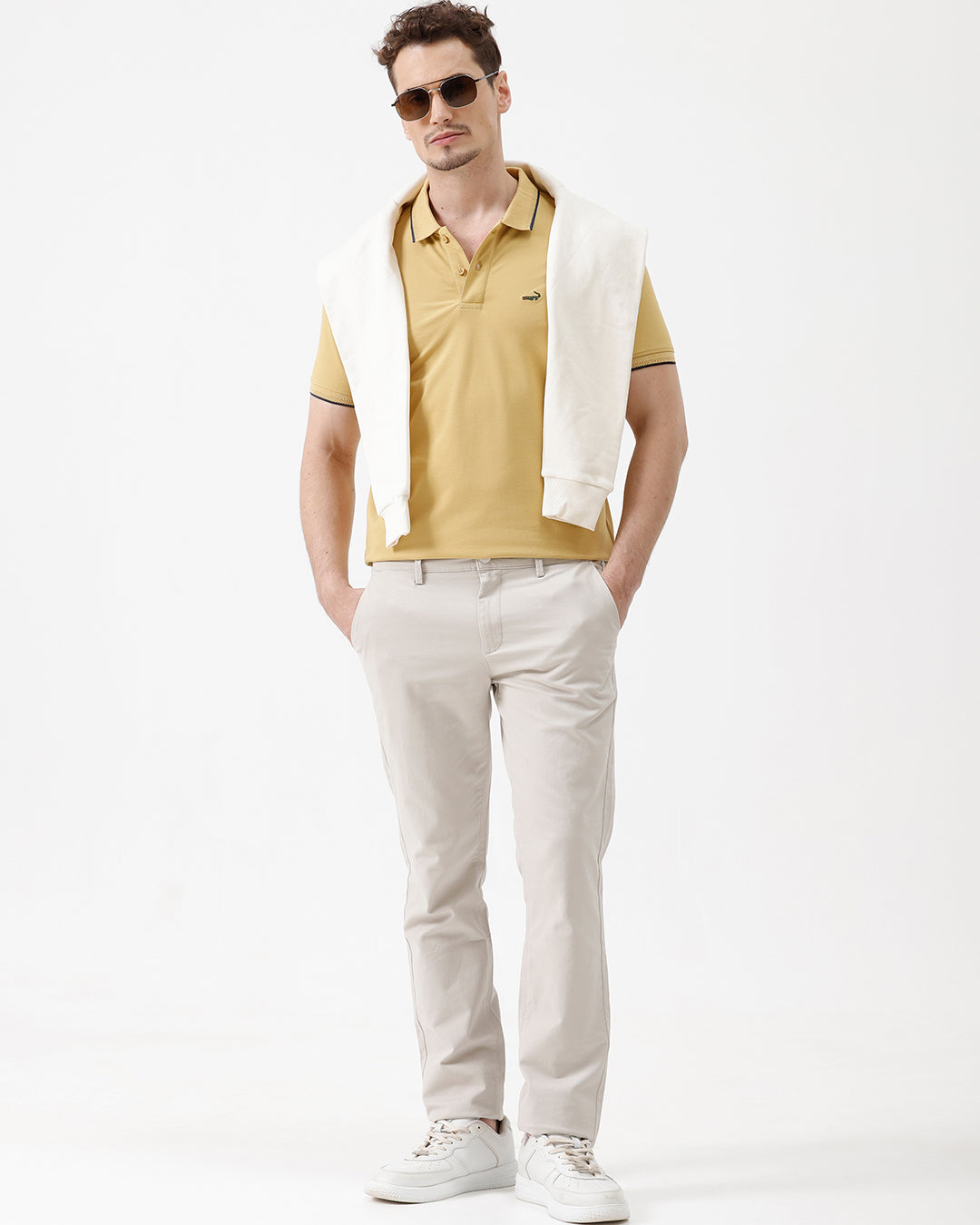 A man wearing a light yellow Lacoste polo shirt with dark piping on the collar and sleeve cuffs, paired with beige trousers, against a plain white studio background.