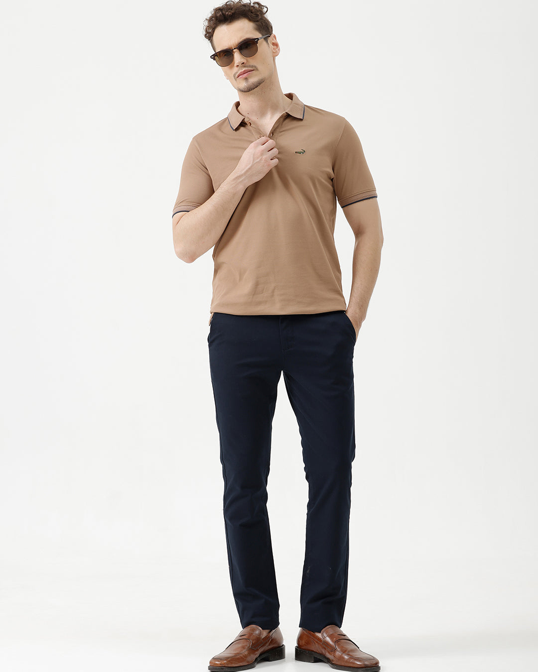A man wearing a tan short-sleeve polo shirt with dark blue piping on the collar and sleeve cuffs, paired with dark navy pants, against a plain light gray background.