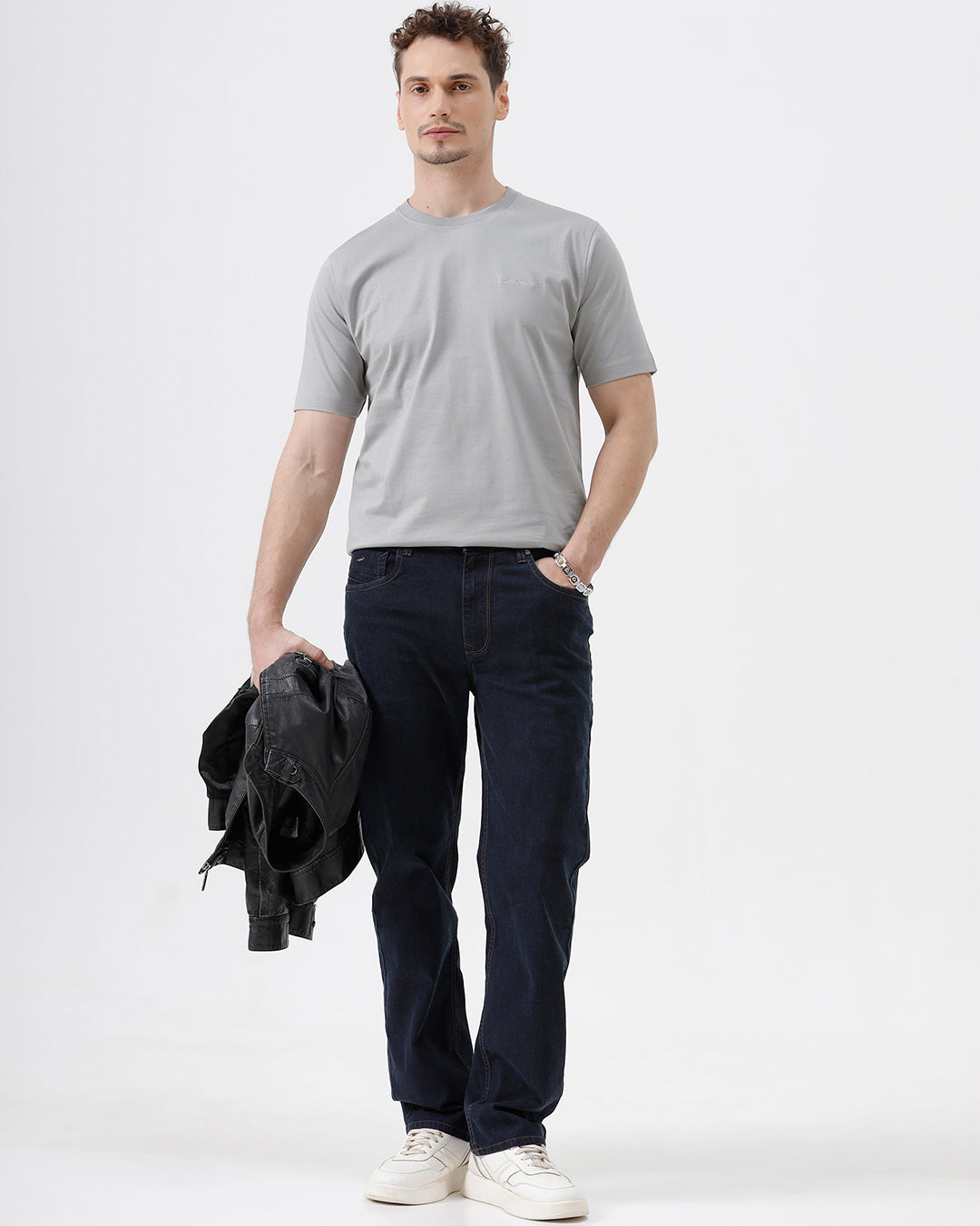 A person wearing dark navy blue straight-leg jeans, a light gray t-shirt, and white sneakers, standing against a plain white studio background.