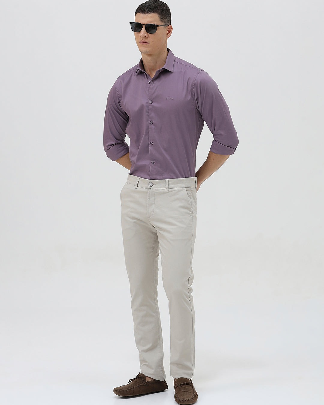 A man wearing a purple long-sleeve button-down shirt with rolled-up sleeves and light beige trousers, standing against a plain light gray background.