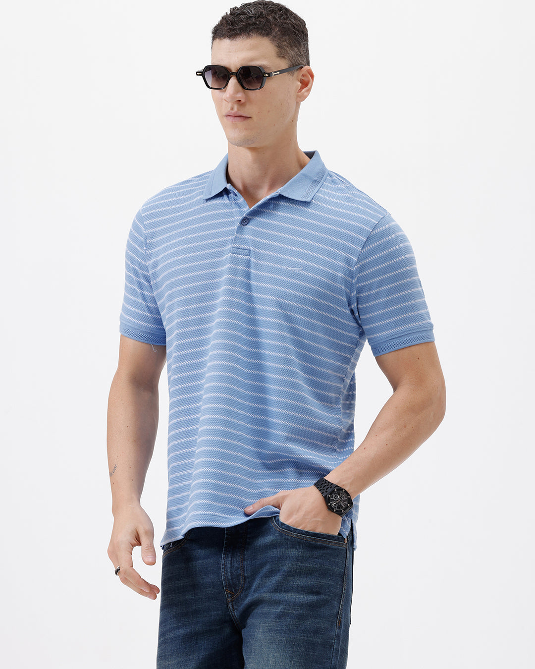 A man wearing a light blue and white striped short-sleeve polo shirt with dark blue jeans, standing against a plain light gray background.