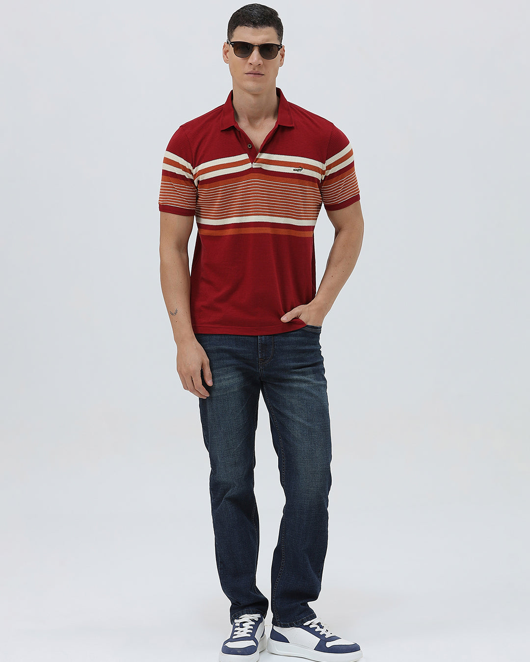 A man wearing a red short-sleeve polo shirt with horizontal cream and rust-orange stripes across the chest and sleeves, paired with dark blue jeans and black sunglasses, against a light gray studio background.