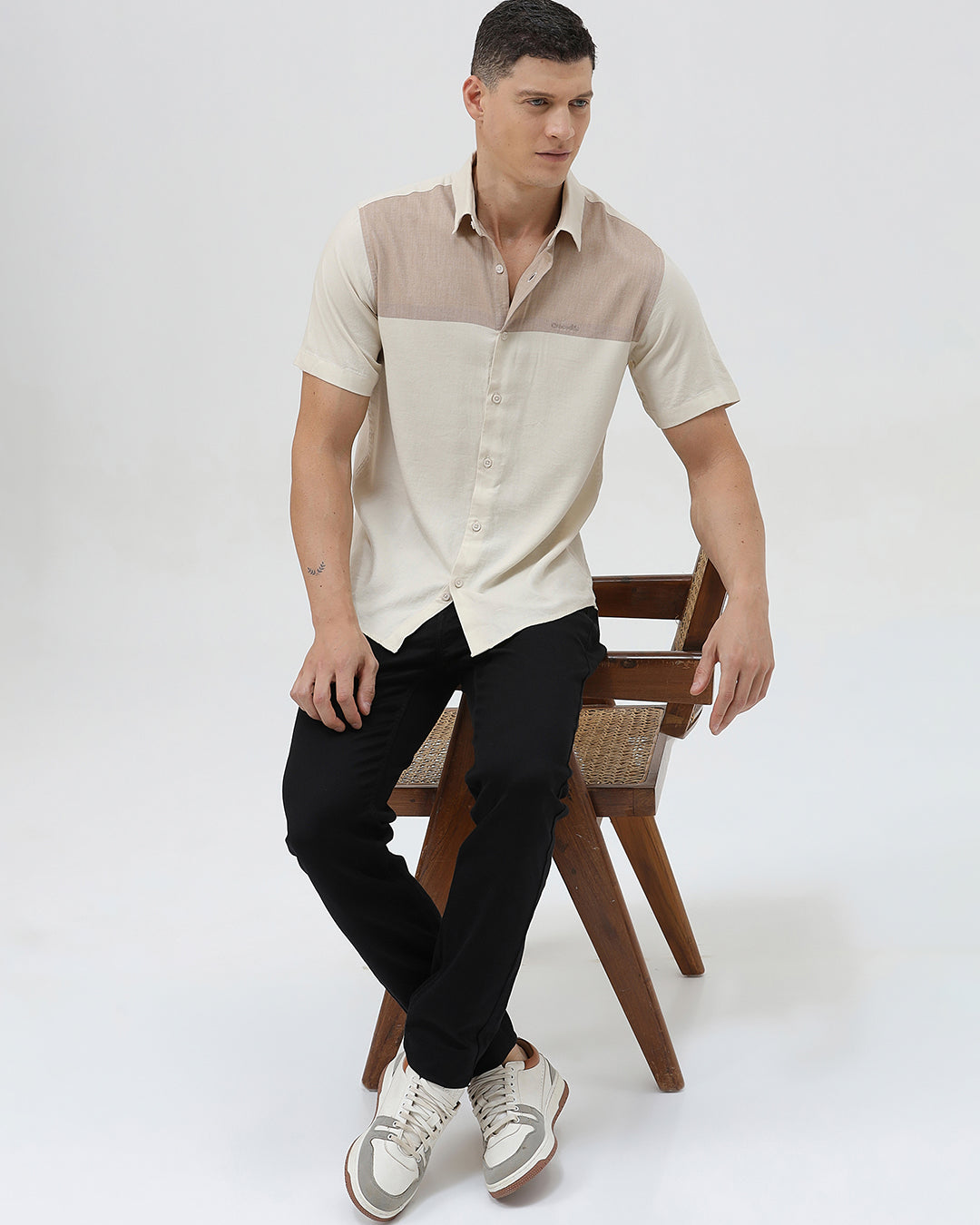 A man wearing a short-sleeve linen button-up shirt with a color-blocked design in beige and cream, paired with black pants, against a plain white studio background.