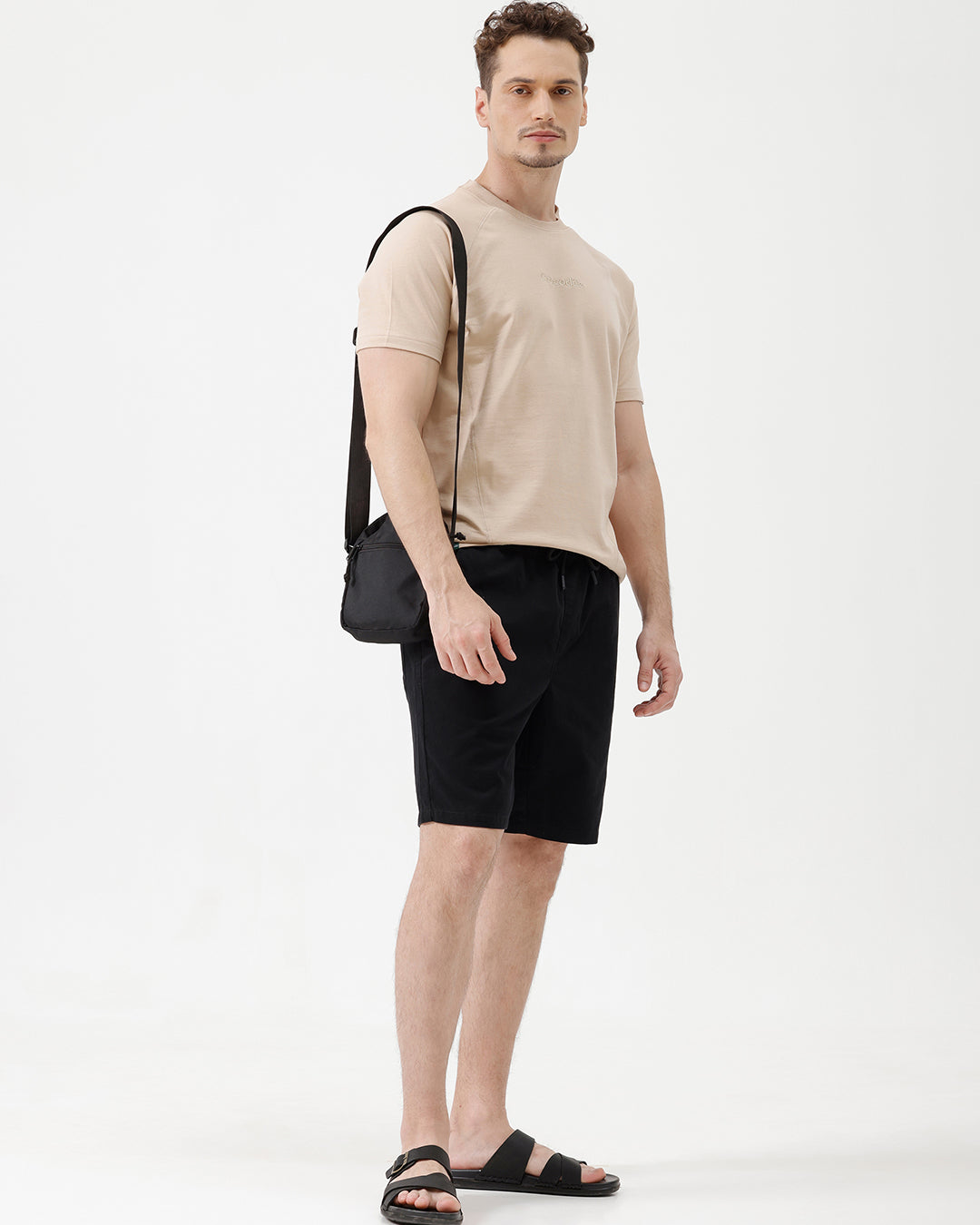 A man wearing a light beige short-sleeve t-shirt with a subtle tonal logo on the chest, paired with black pants, standing against a plain white studio background.