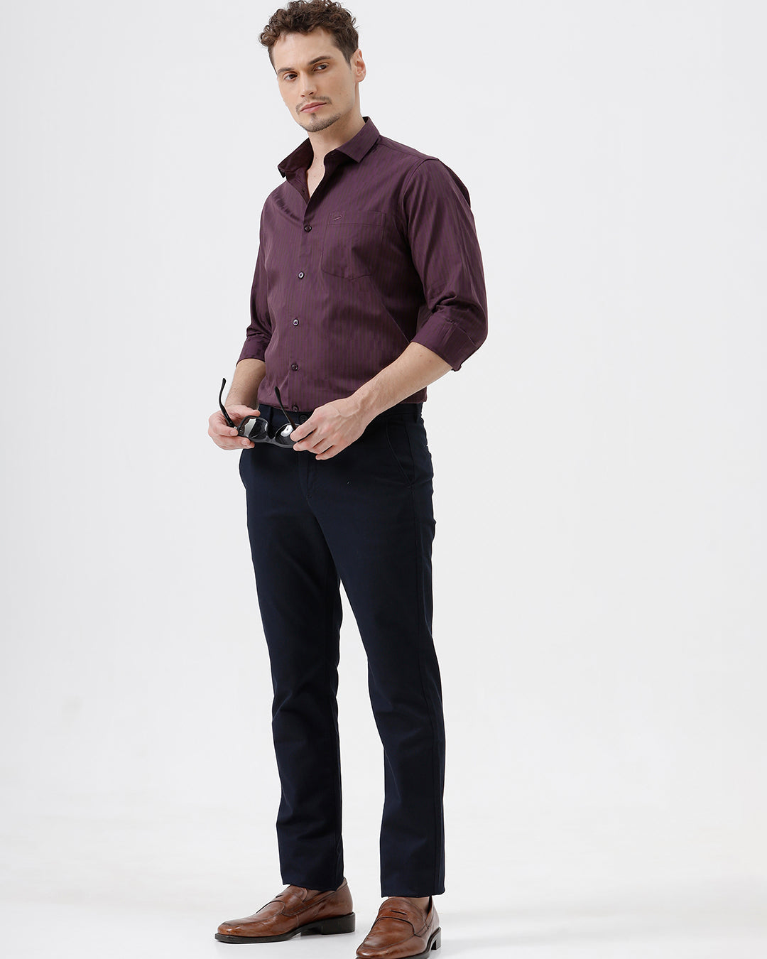A man wearing a dark purple long-sleeve button-down shirt with subtle vertical stripes and dark navy trousers, standing against a light gray background.