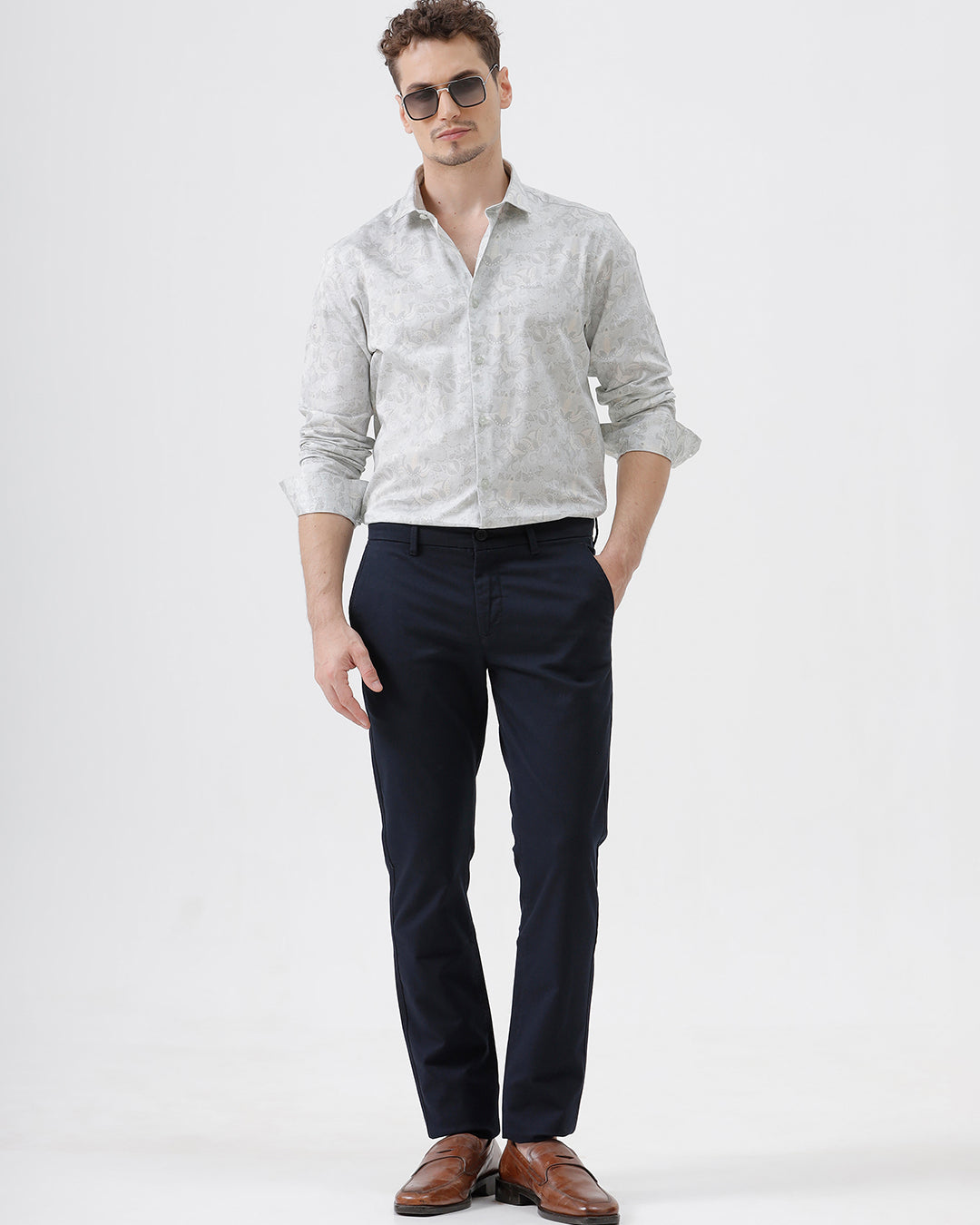 A man wearing dark navy blue straight-leg trousers, a light-colored patterned shirt, and brown leather loafers, standing against a plain white studio background.