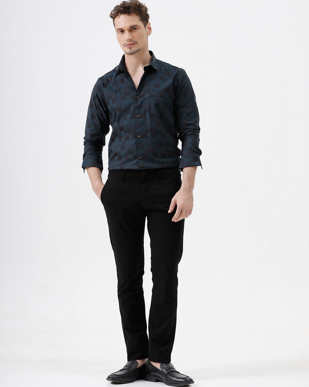A man wearing a dark navy blue patterned long-sleeve button-up shirt and black trousers, standing against a plain white studio background.