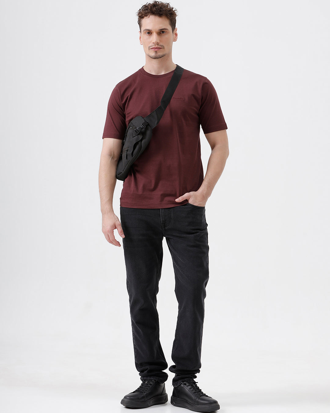 A man wearing a short-sleeve crewneck t-shirt in maroon, paired with dark jeans, standing against a plain light gray studio background.