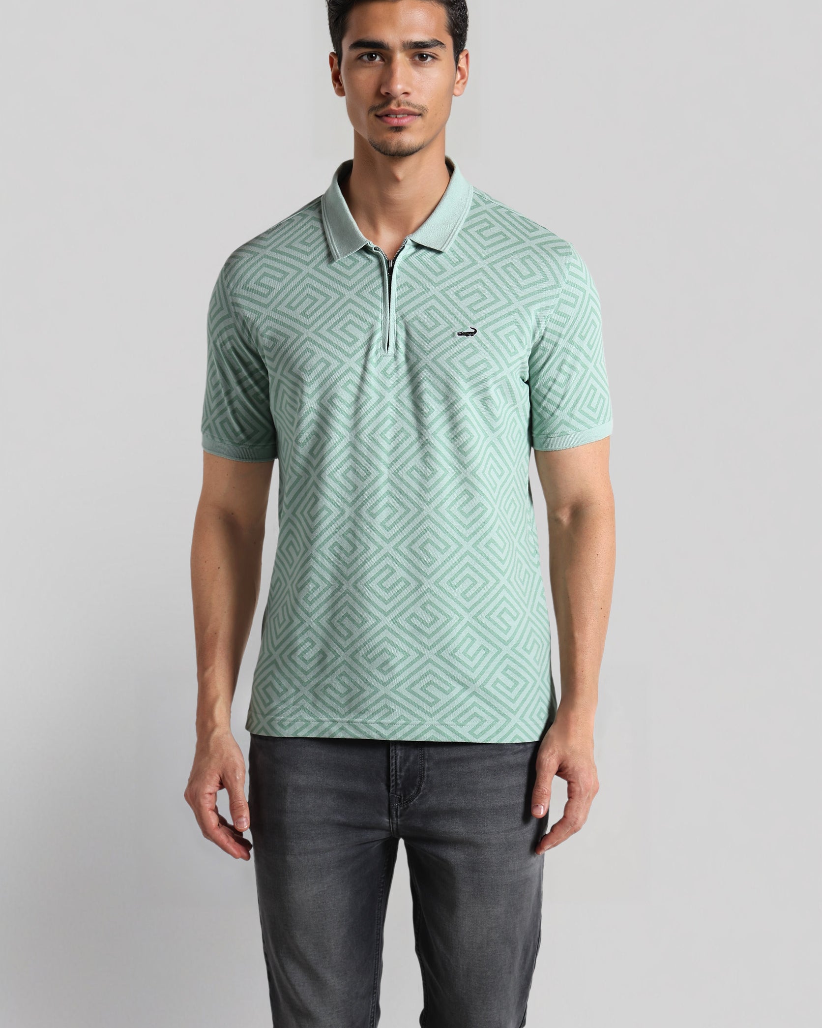 Man wearing a light green patterned polo shirt and dark jeans on a gray background