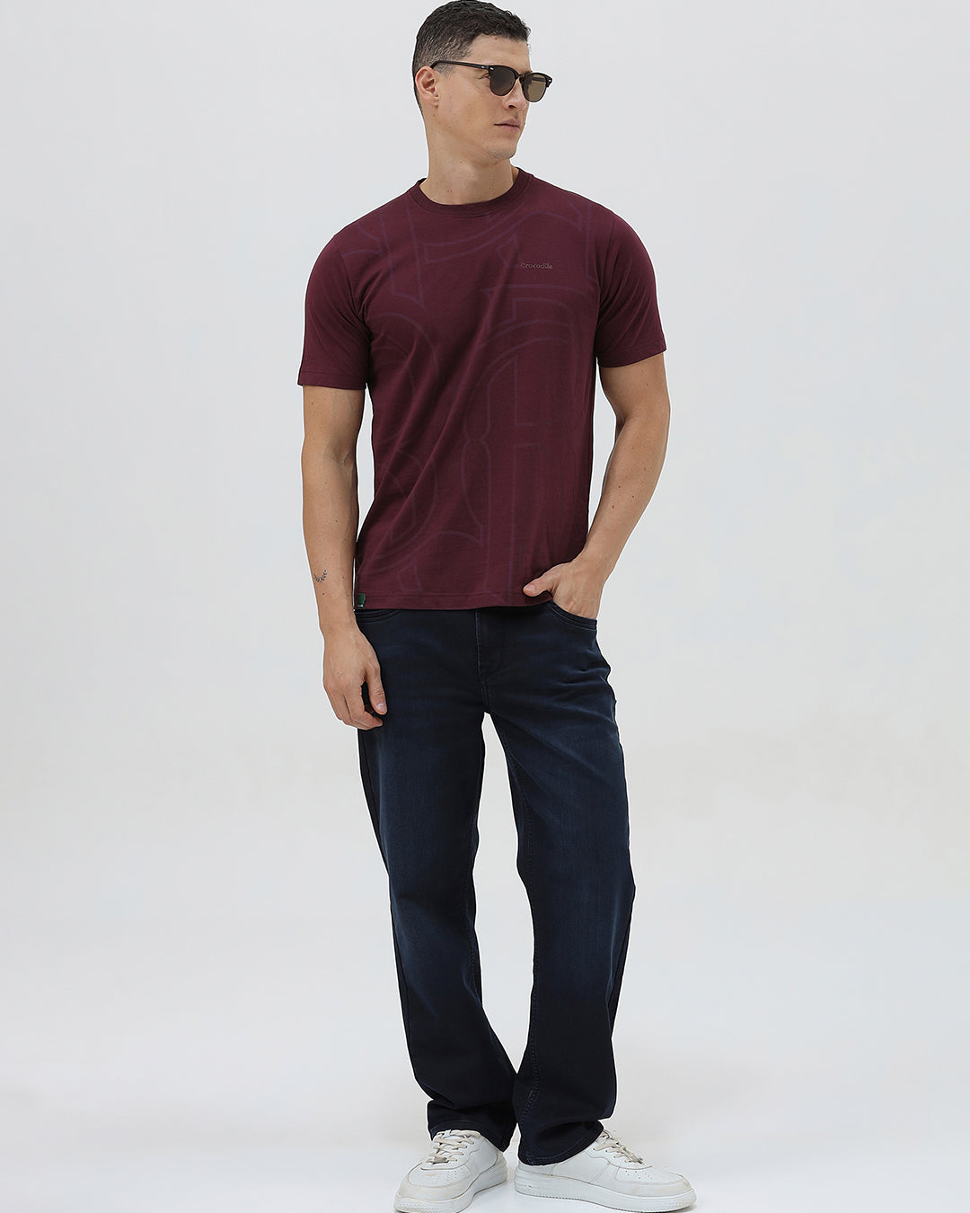 A man wearing a short-sleeve crewneck t-shirt in maroon with a subtle tonal graphic pattern, paired with dark blue jeans, against a light gray studio background.