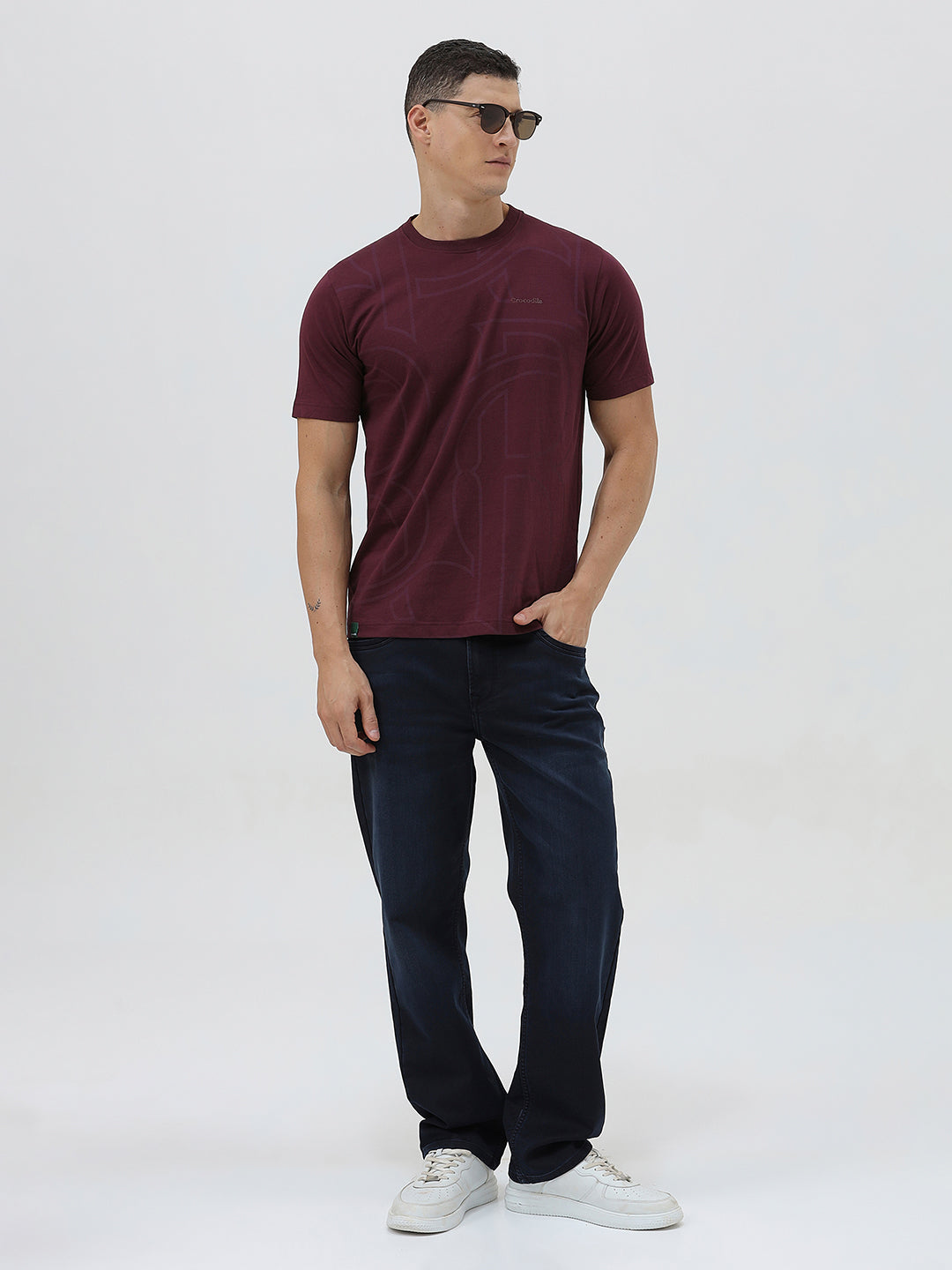 A man wearing a short-sleeve crewneck t-shirt in maroon with a subtle tonal graphic pattern, paired with dark blue jeans, against a light gray studio background.