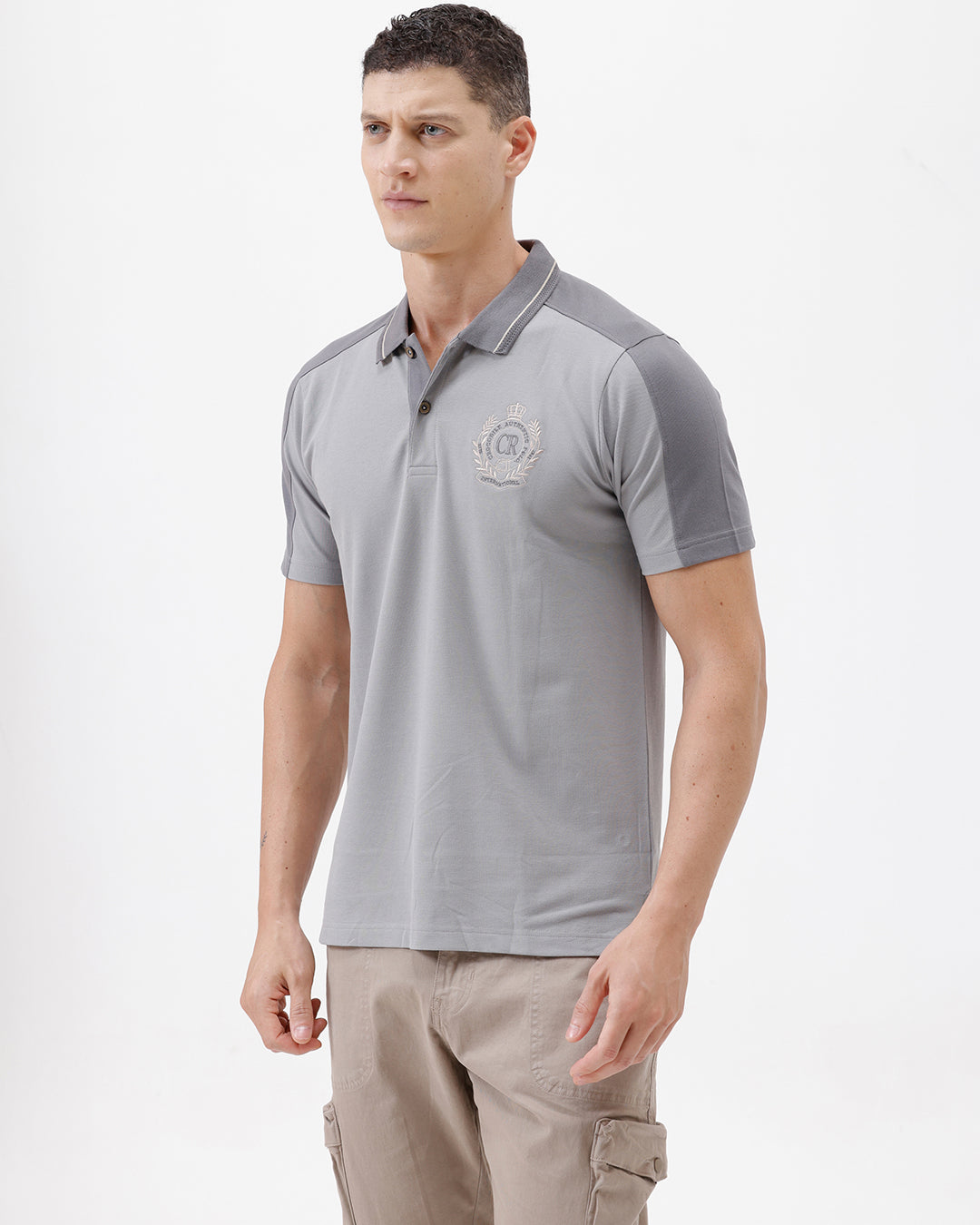 A man wearing a light gray short-sleeve polo shirt with darker gray shoulder panels and a small crest logo on the left chest, paired with khaki cargo pants, against a plain light gray studio background.