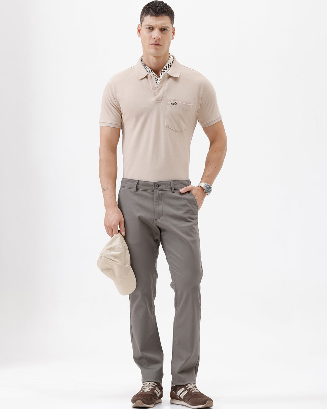 A man wearing a light beige short-sleeve polo shirt with a small crocodile logo on the chest pocket, paired with gray trousers, photographed against a plain white studio background.