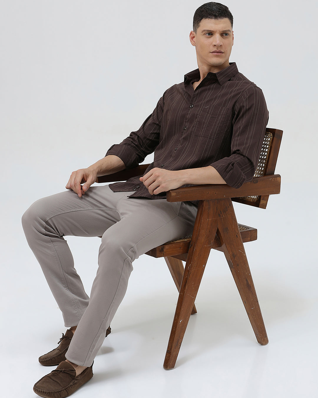 A man wearing a dark brown long-sleeve button-up shirt with thin vertical stripes and light gray trousers, standing against a plain light gray background.