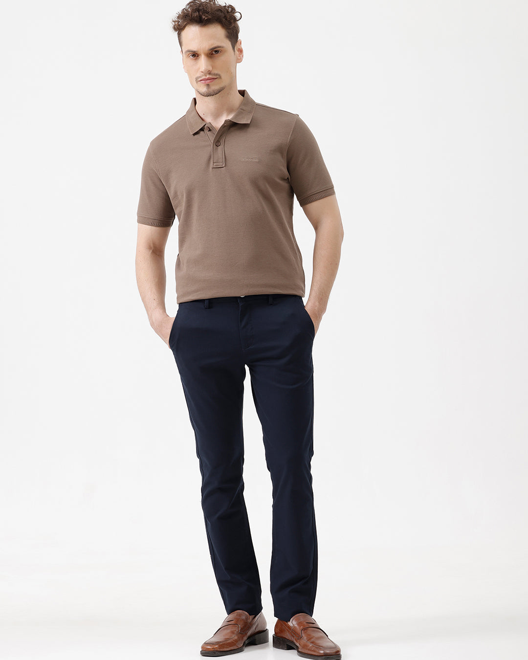 A man wearing a brown short-sleeve polo shirt with dark navy pants, standing against a plain light gray studio background.