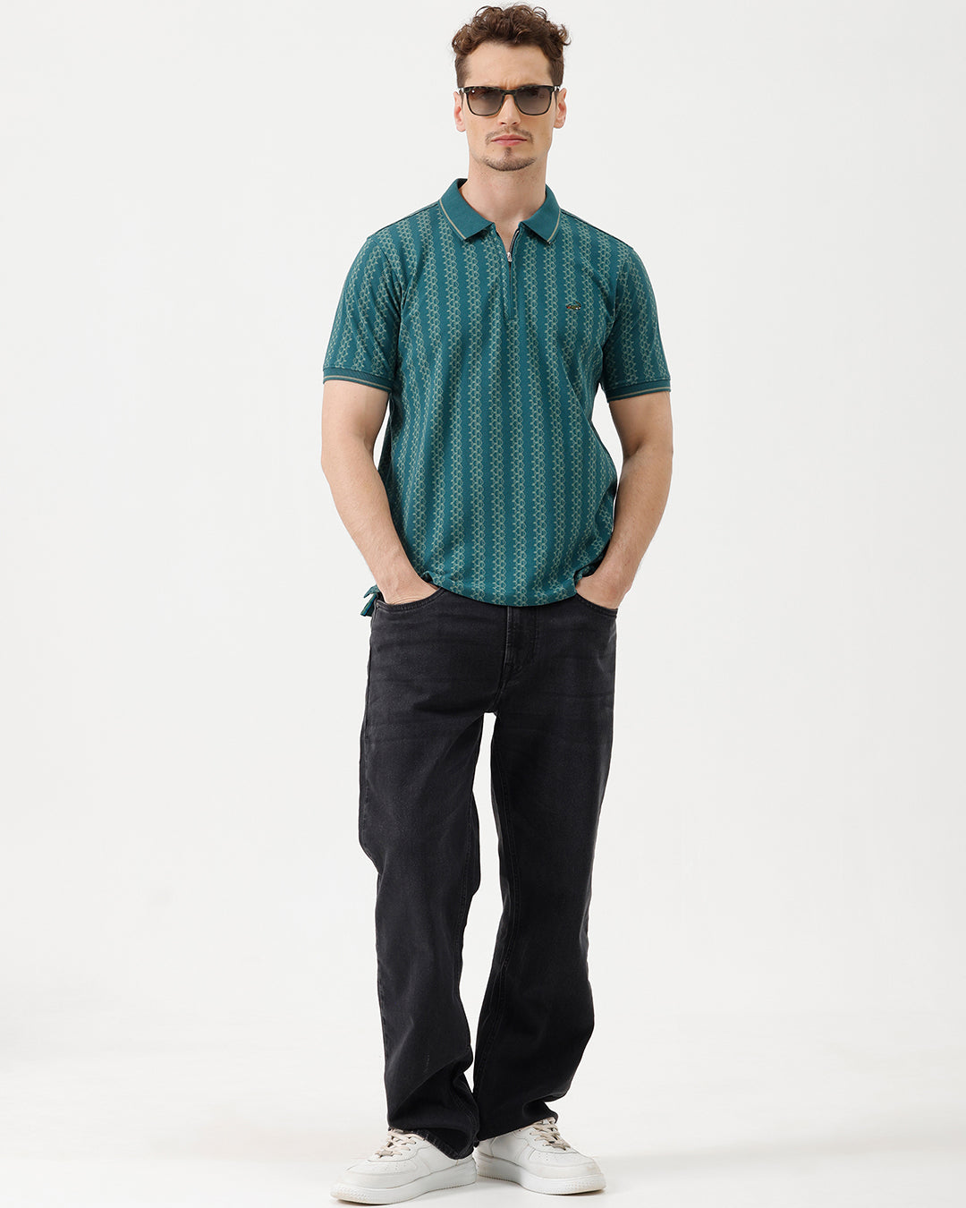 A man wearing a teal green short-sleeve polo shirt with a white geometric pattern, paired with black jeans, standing against a plain white studio background.