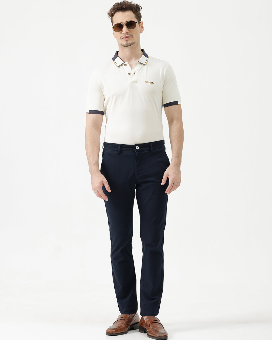 A man wearing dark navy blue slim-fit trousers, a cream-colored shirt, and brown leather loafers, standing against a plain white studio background.