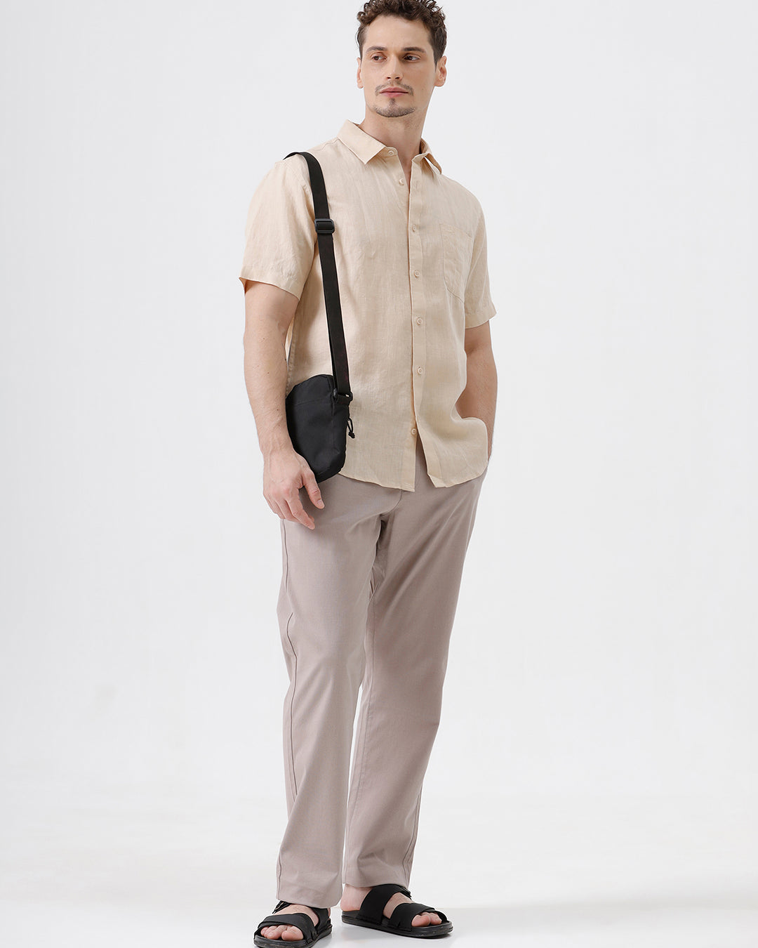 A man wearing a light beige short-sleeve linen button-up shirt and taupe trousers, standing against a plain white studio background.