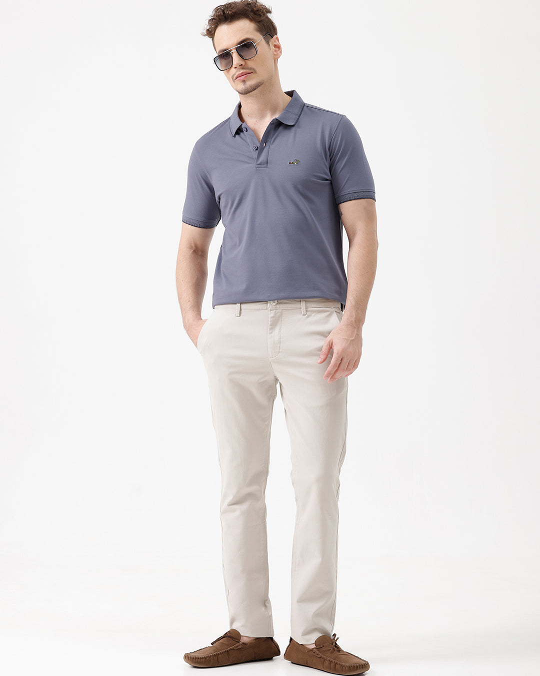 A person wearing light beige chino pants, a dark blue-gray t-shirt, and brown suede moccasin shoes, standing against a plain white studio background.
