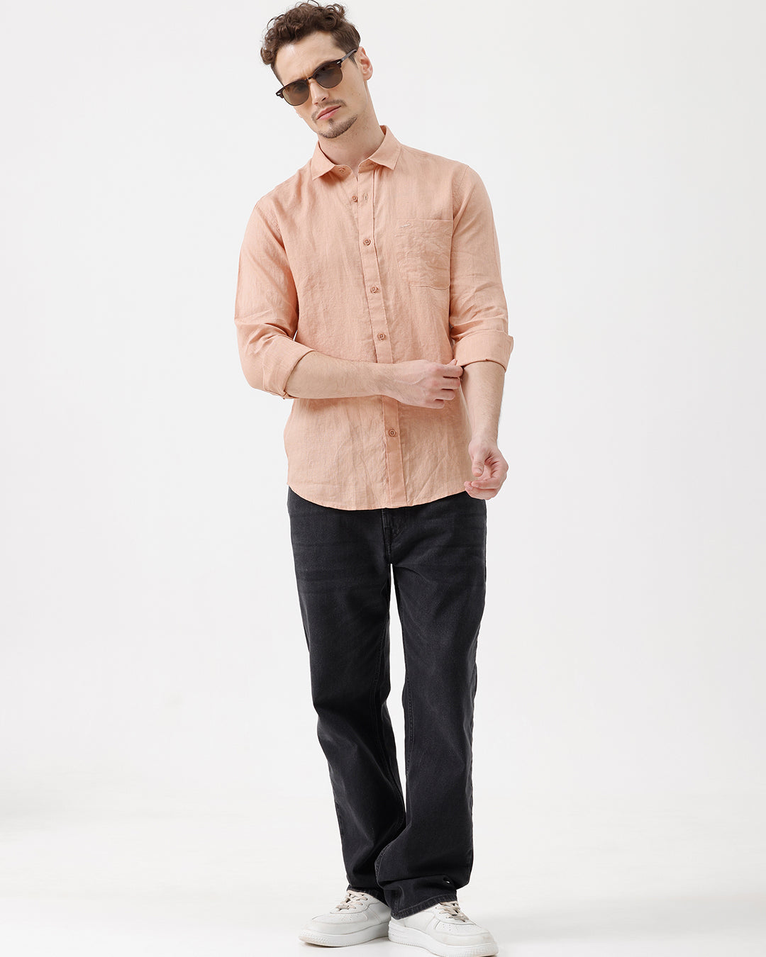 A man wearing a light peach-colored linen button-down shirt with dark jeans, standing against a plain white studio background.