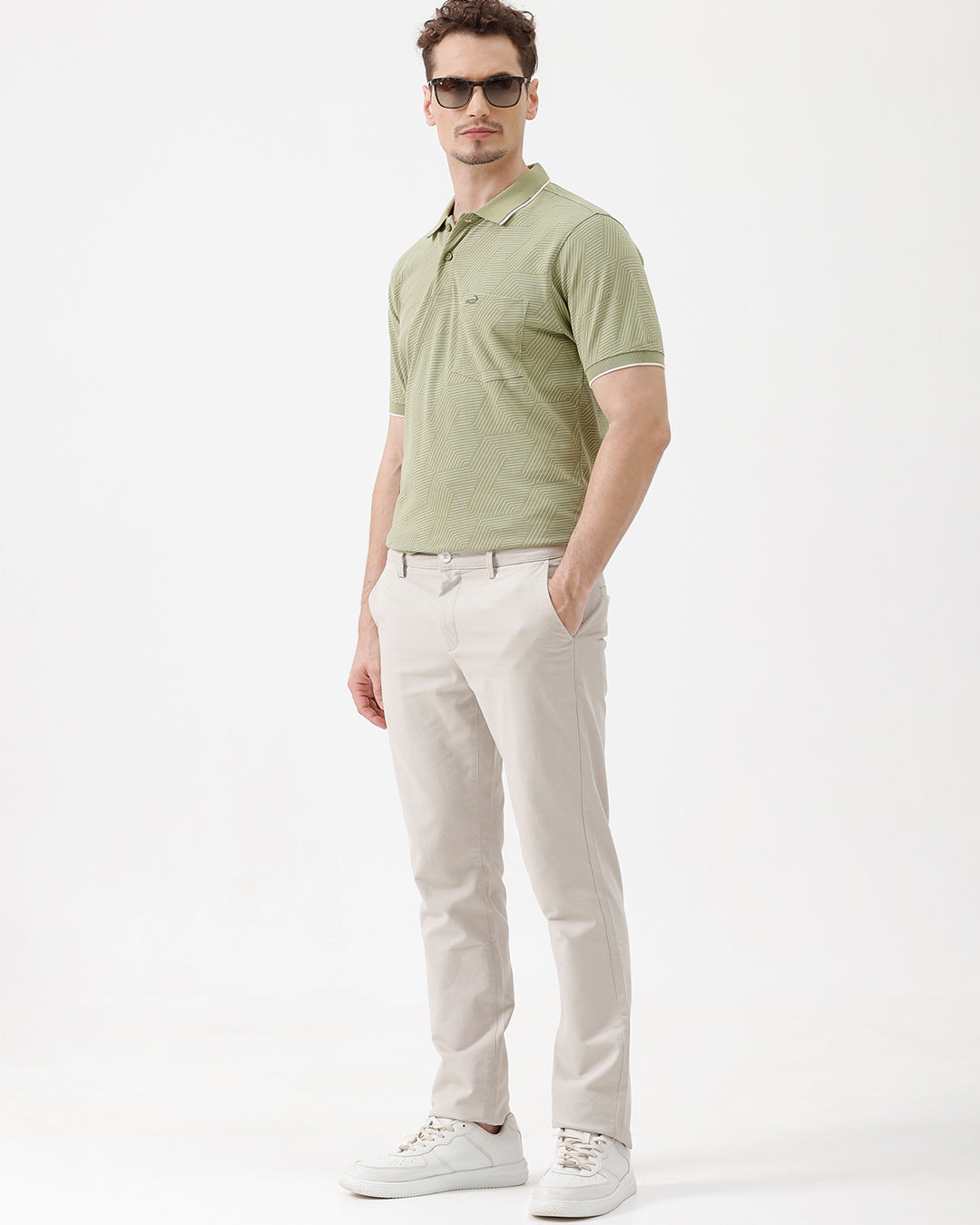 A man wearing a light sage green short-sleeve polo shirt with a subtle geometric pattern, paired with beige trousers, standing against a plain white studio background.