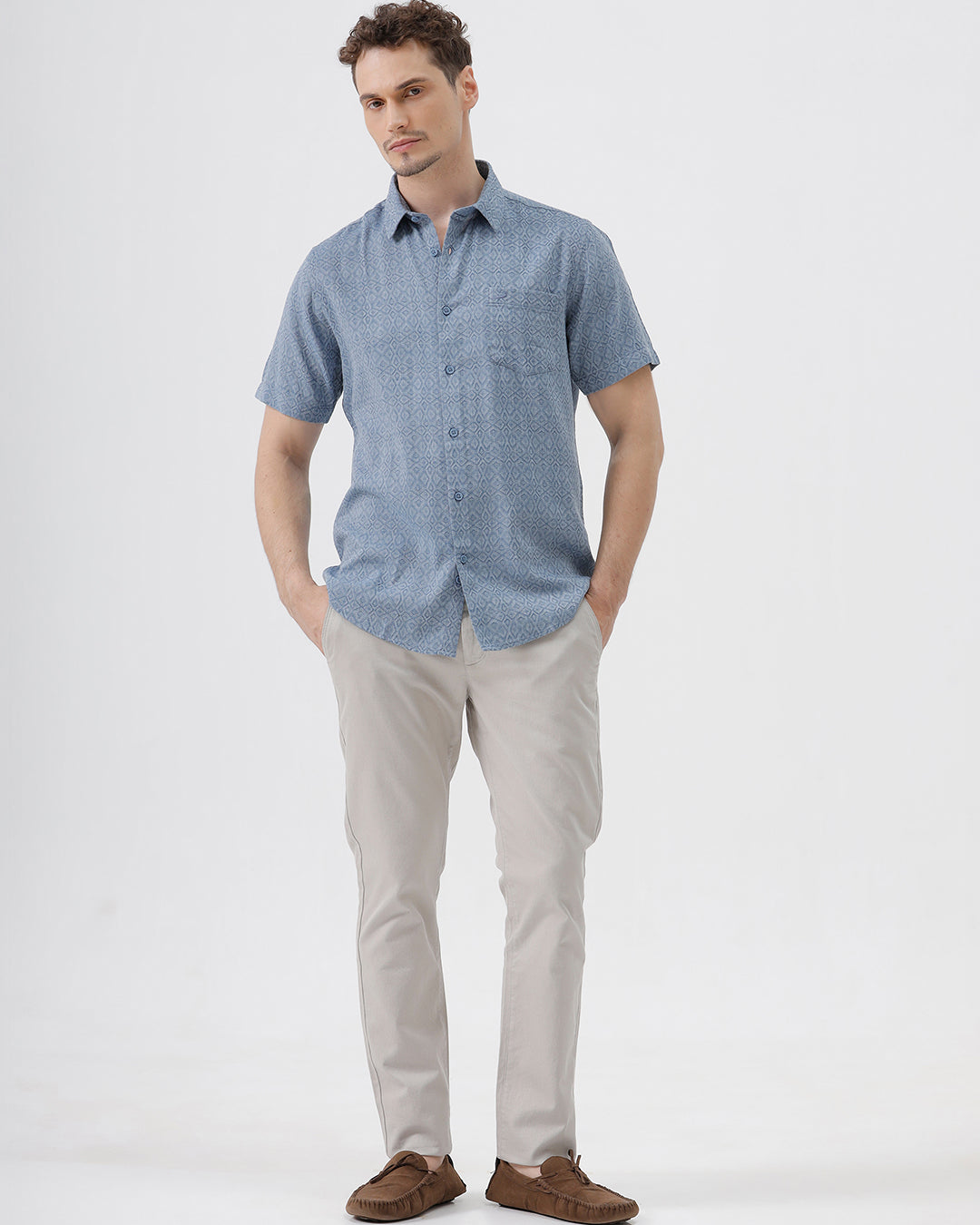 A man wearing a short-sleeve button-up shirt with a fine blue and white geometric pattern, paired with light beige trousers, standing against a plain light gray background.