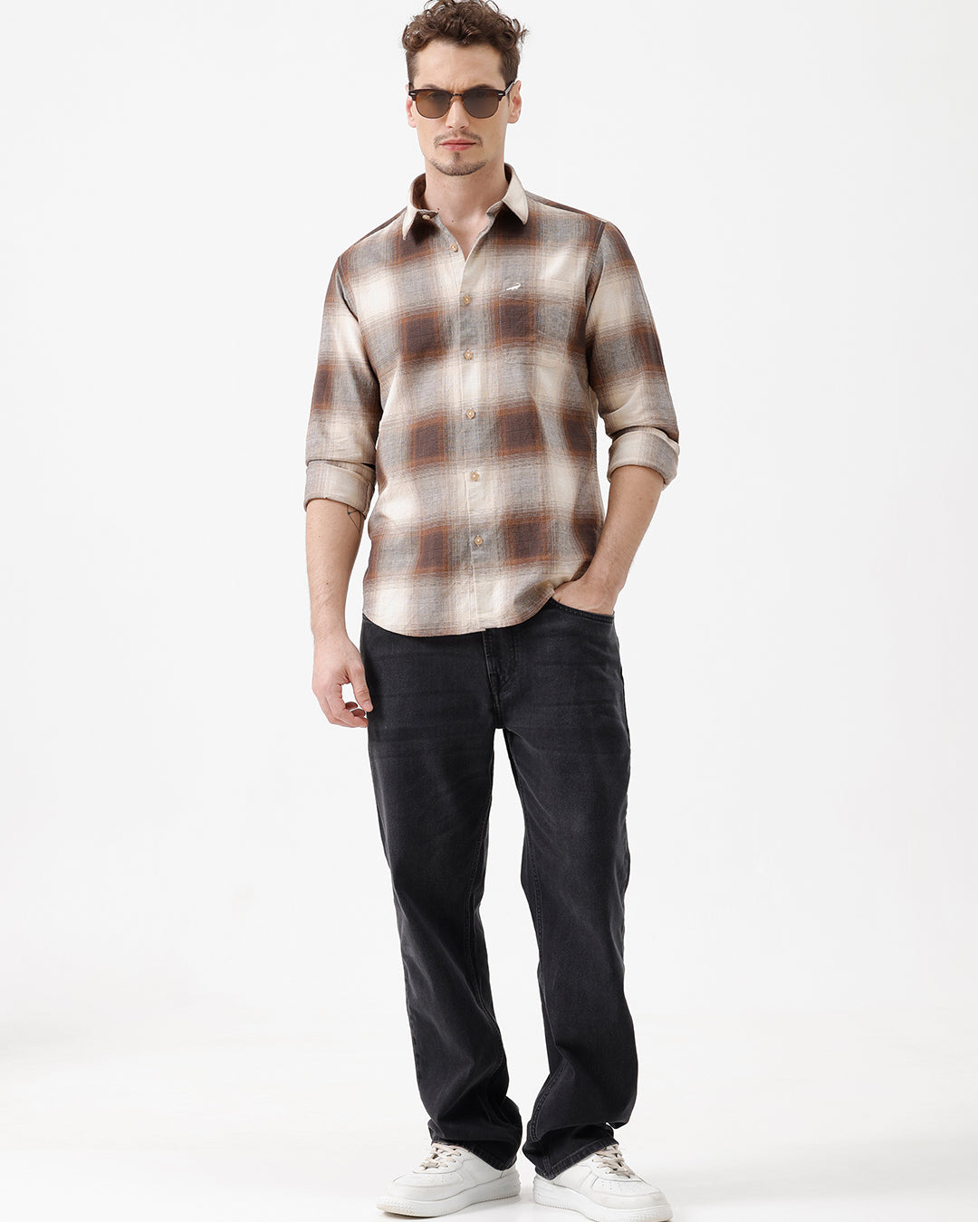 A man wearing a long-sleeve button-up plaid shirt in brown, beige, and gray tones, paired with dark jeans, standing against a plain white studio background.